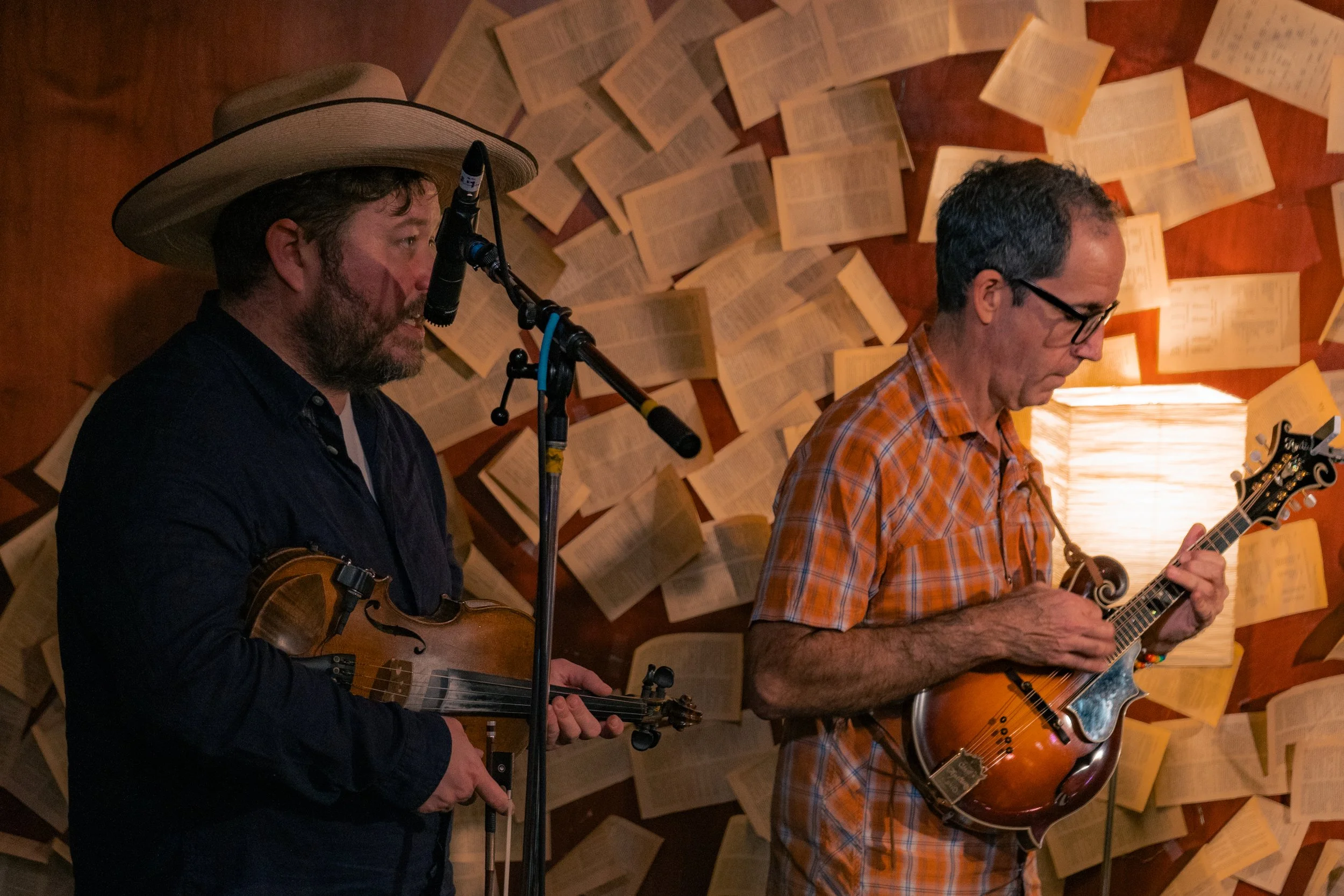 Two men playing stringed instruments, one with a hat, surrounded by open books on a wall and a lamp.
