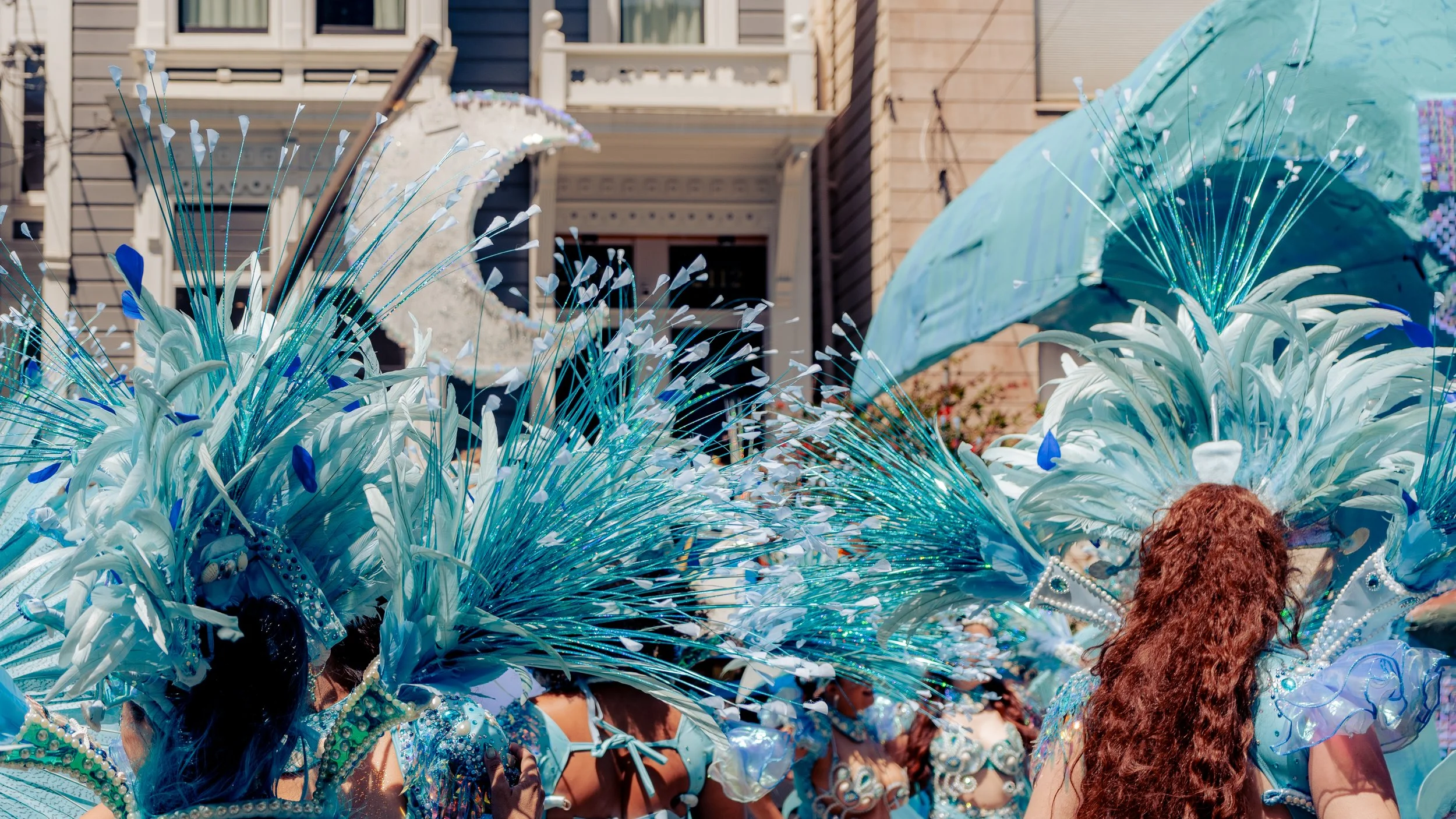 People dressed in elaborate blue costumes with feather and glitter details participate in a parade, with houses in the background.