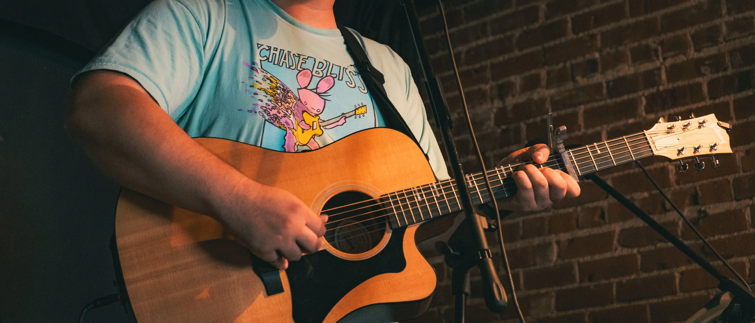 Person playing acoustic guitar with a brick wall in the background, wearing a light blue t-shirt with a colorful graphic of a mouse playing guitar.