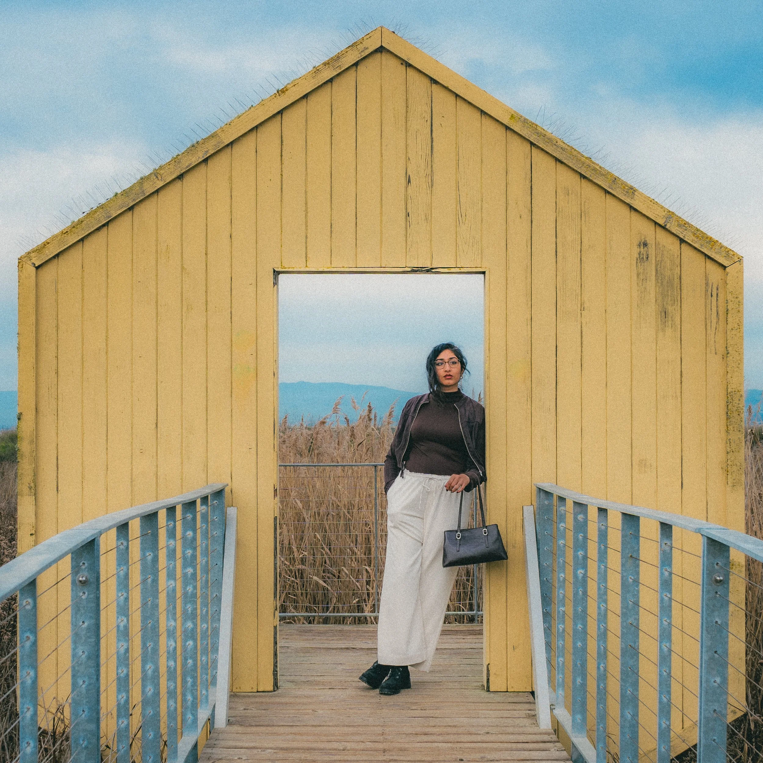 A woman standing in front of a yellow wooden structure on a small bridge, with tall grasses and mountains in the background.