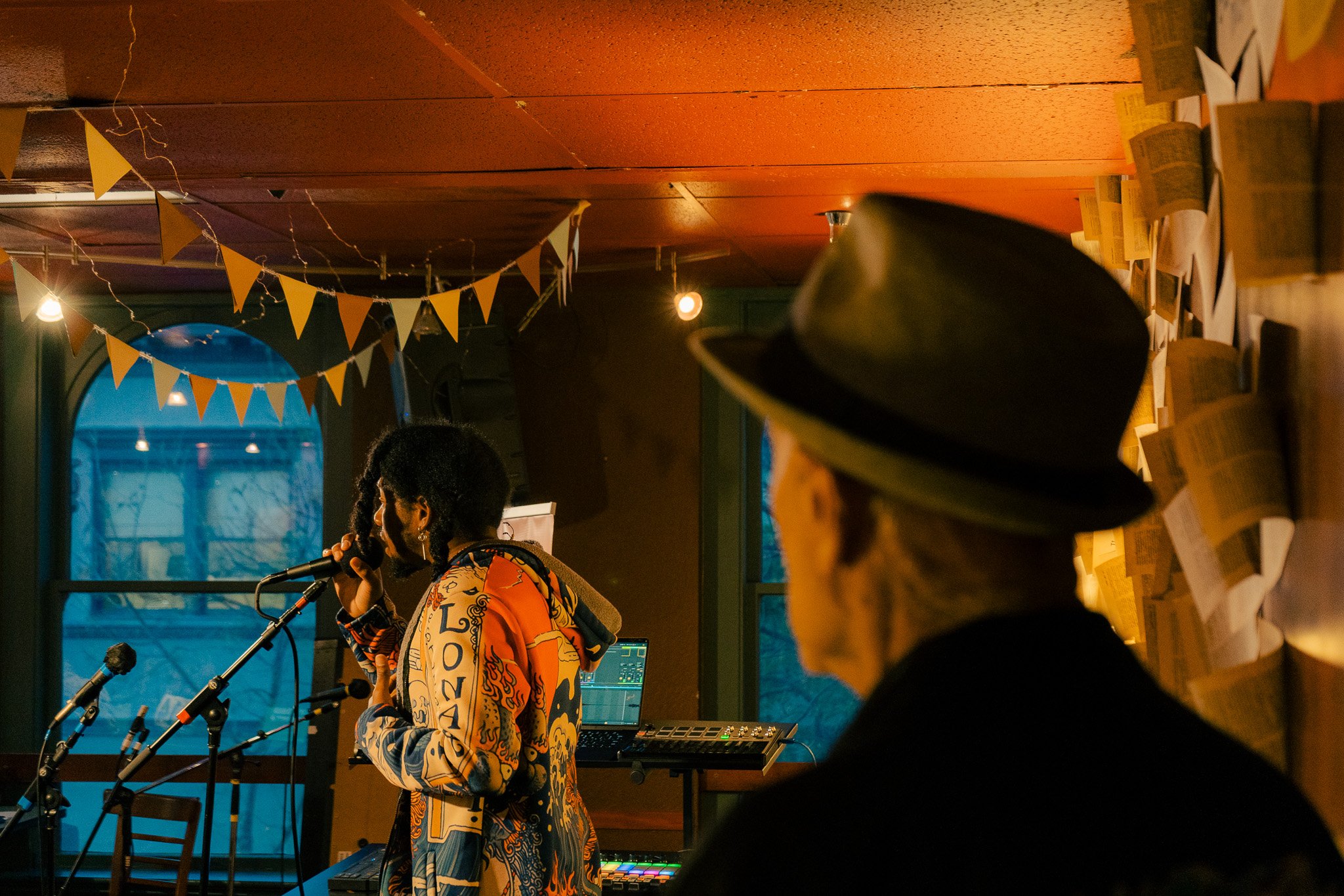 A woman singing into a microphone with an electronic music setup, in a cozy room decorated with yellow triangular flags hanging from the ceiling and papers pasted on the wall, while a person wearing a wide-brimmed hat observes.