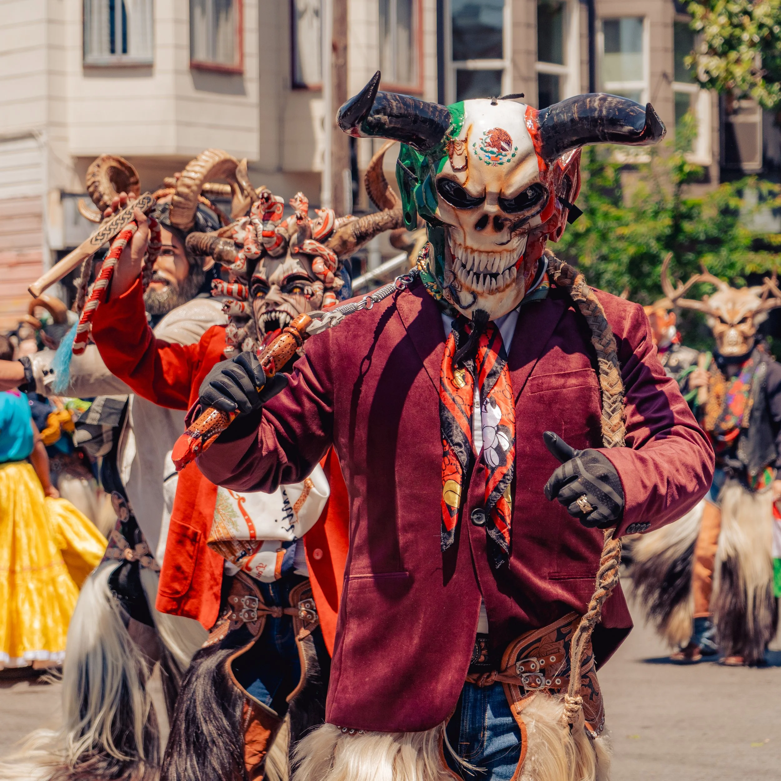 People dressed in elaborate costumes with masks, horns, and accessories participating in a cultural outdoor festival or parade, with buildings and trees in the background.