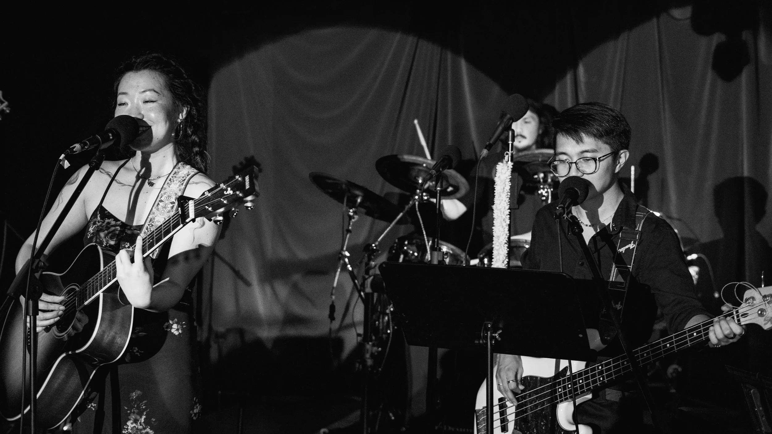 Three musicians performing on stage, two women playing guitars and a man on drums, with microphones and musical equipment.