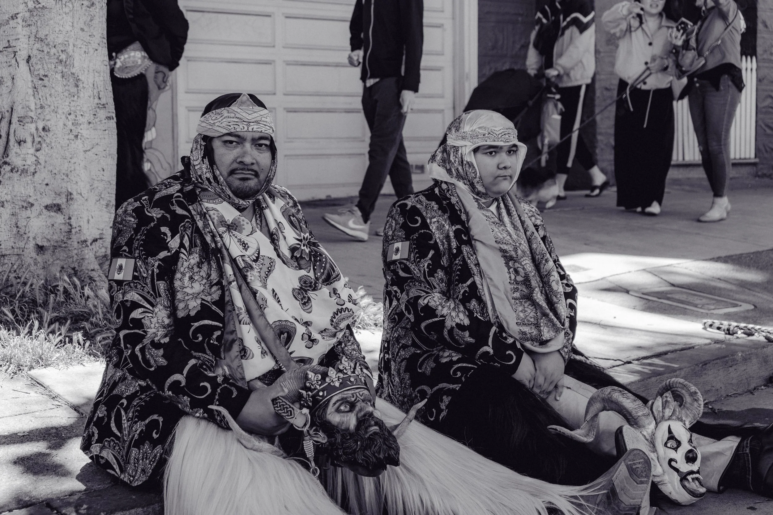 Two individuals dressed in traditional indigenous costumes sitting on the sidewalk, with masks and decorative clothing, while a group of people walk past in the background.