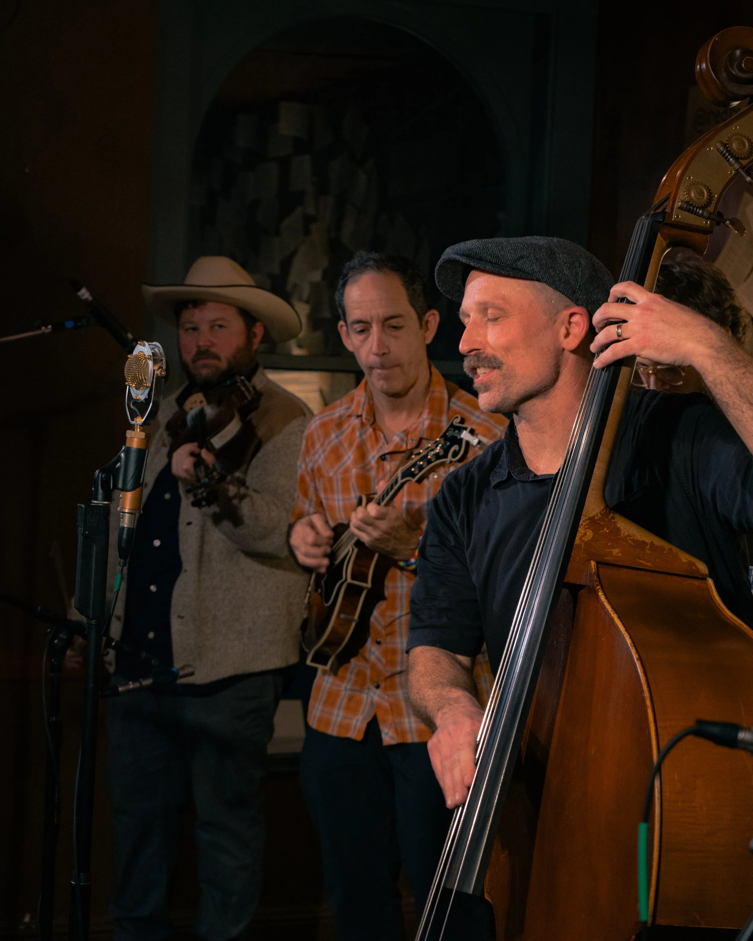 Three musicians performing in a cozy, dimly lit setting, one playing cello, one with a guitar, and one with a fiddle, with a vintage microphone nearby.