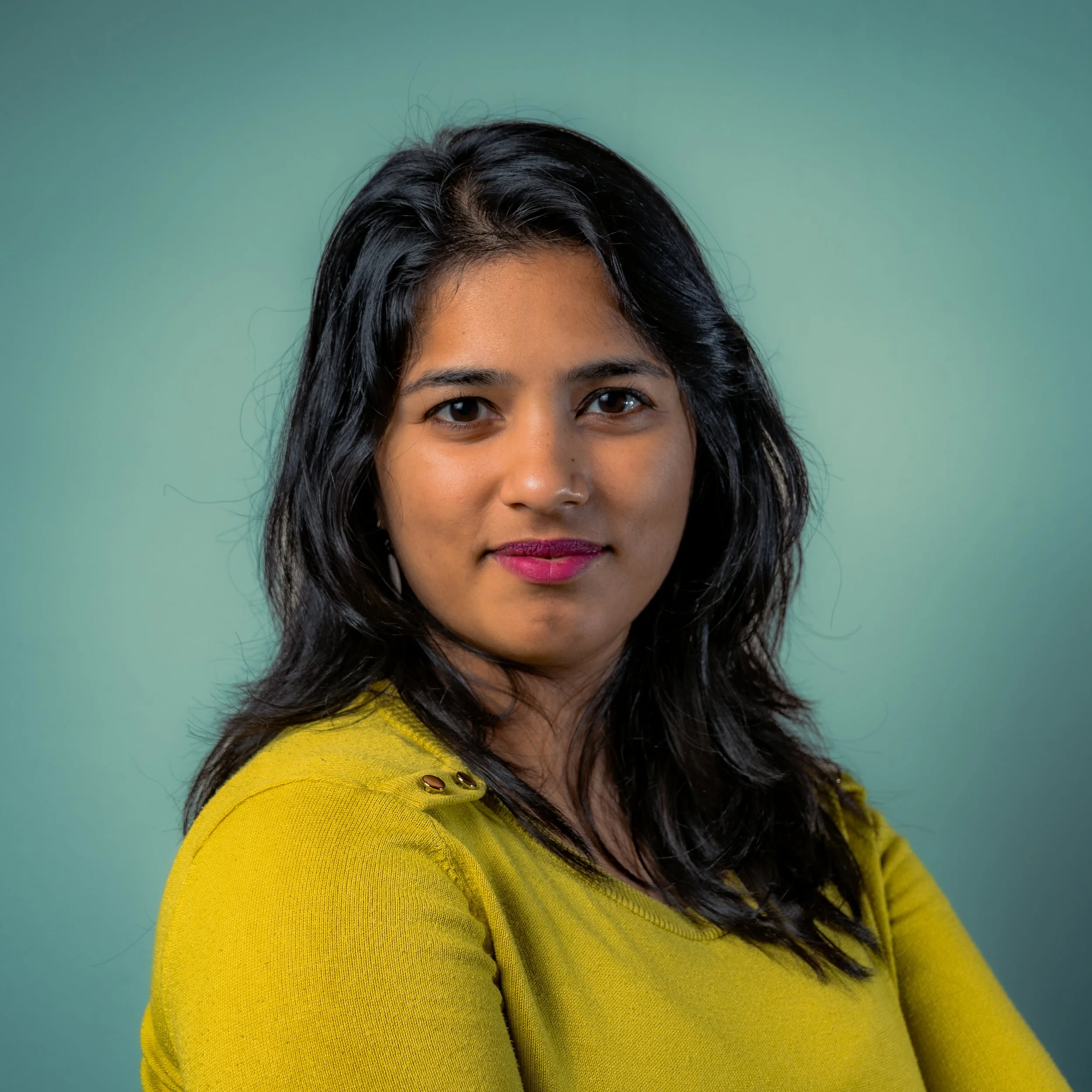 A woman with dark, wavy hair and medium skin tone, wearing a yellow top, standing against a green gradient background.