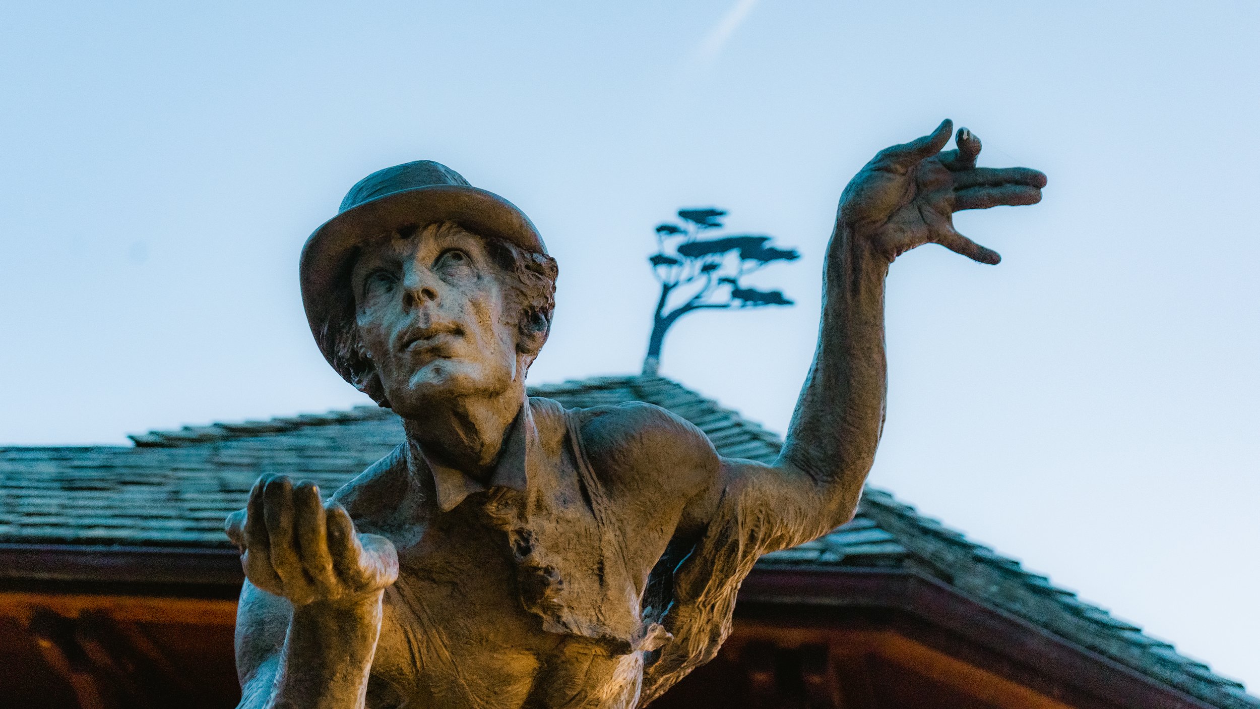 Bronze statue of a woman with a hat, gesturing with her right hand and looking upward, with a tree and roof in the background.