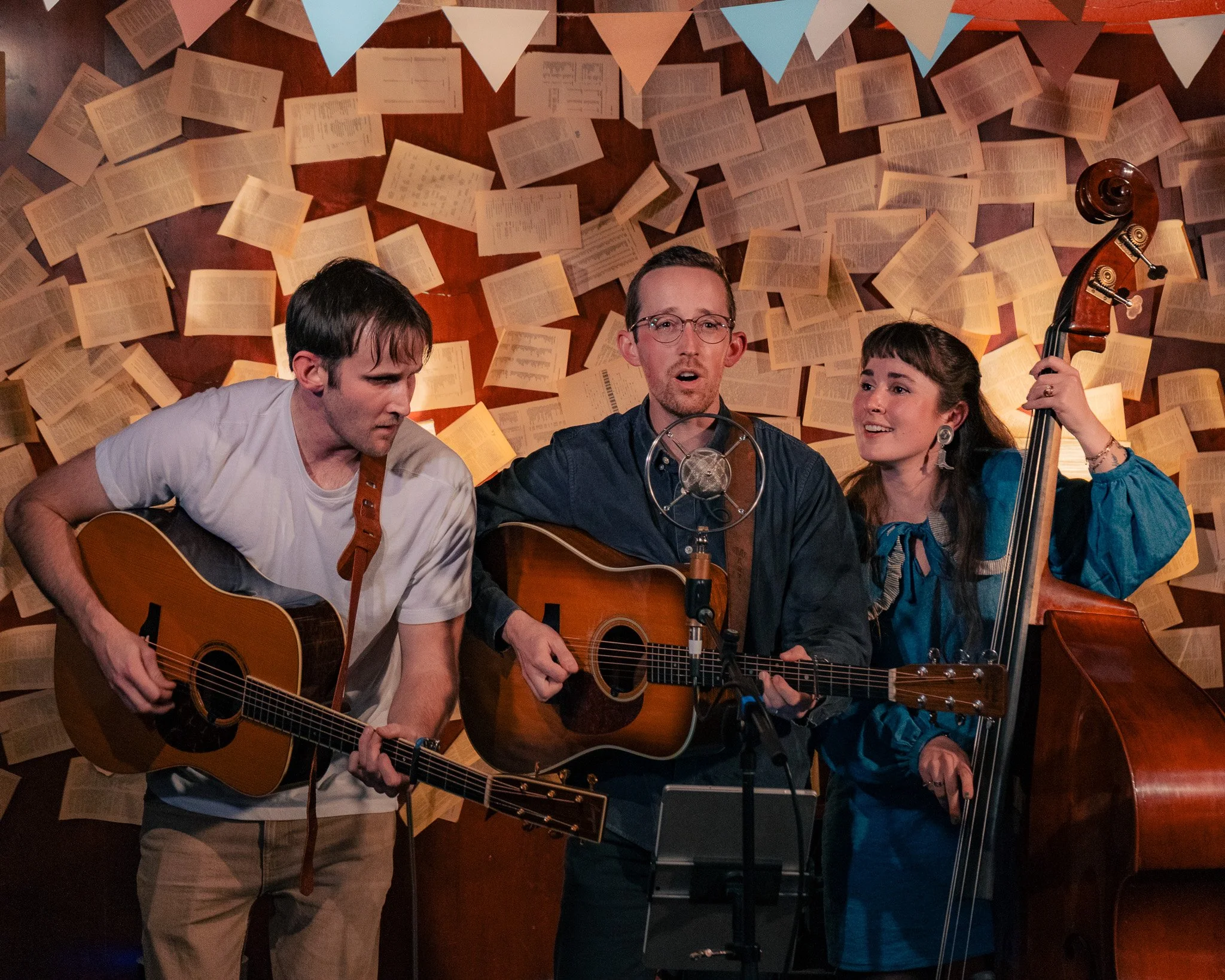 Three musicians performing in a cozy, acoustic setting decorated with hanging papers and triangular banners. The man in the middle is singing and playing an acoustic guitar, the man on the left is playing a guitar, and the woman on the right is playi