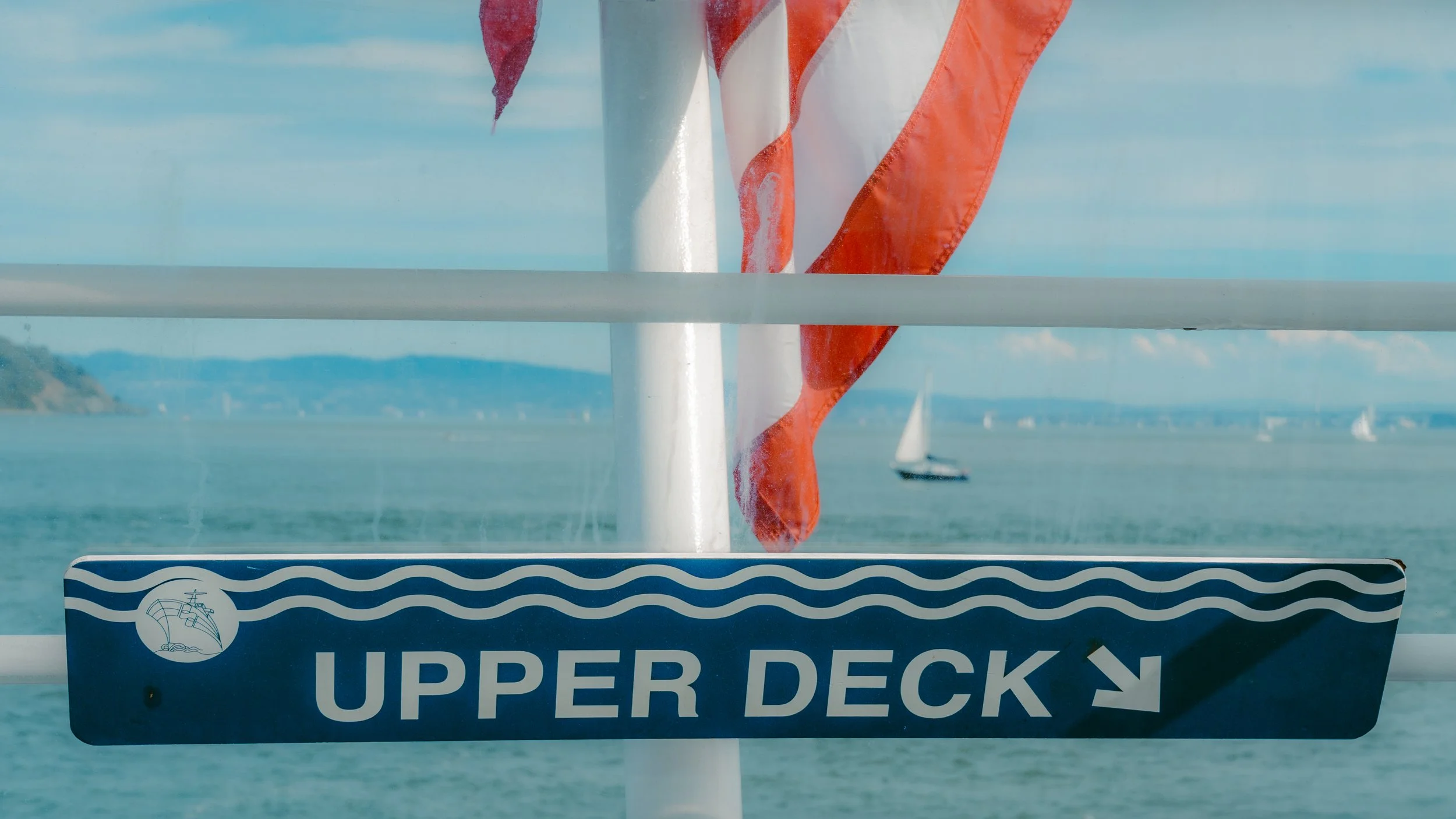 A sign on a boat indicates the upper deck, with a background view of the ocean, sailboats, and distant shoreline.