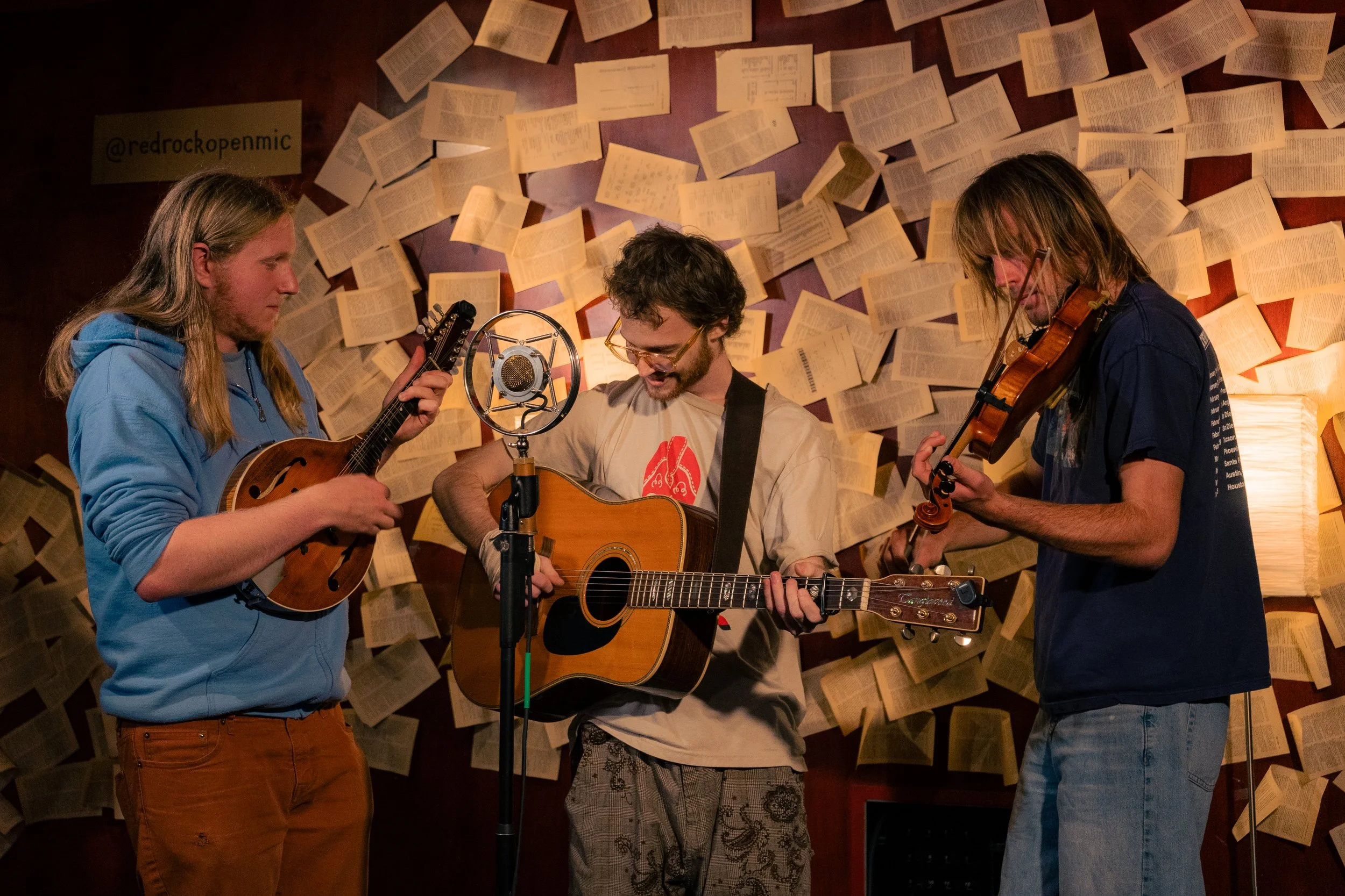 Three young men playing string instruments and singing into a vintage microphone in a cozy room with a backdrop of open books on the wall.