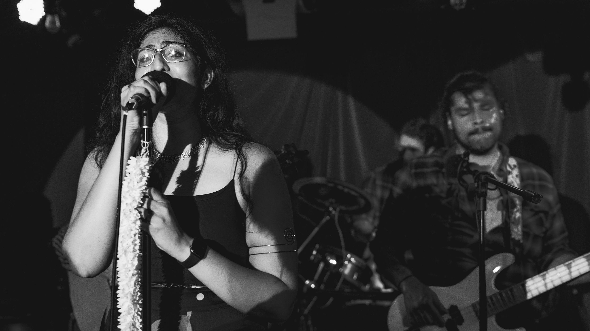 A woman singing into a microphone on stage, with band members playing guitars and drums in the background, in a dimly lit setting.