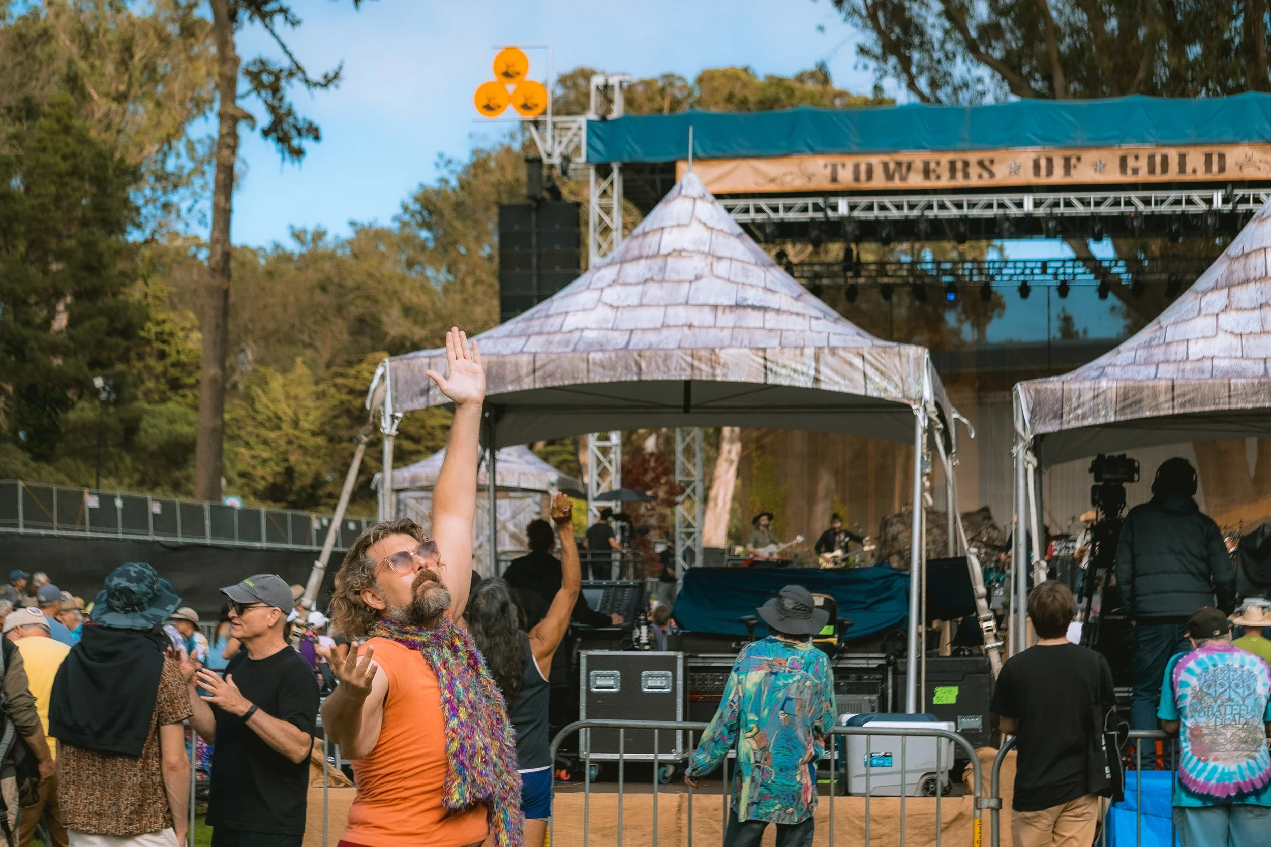 People attending an outdoor concert at the TOWERS of GOLD stage, some with hands raised, in front of a stage with performers playing, surrounded by trees.