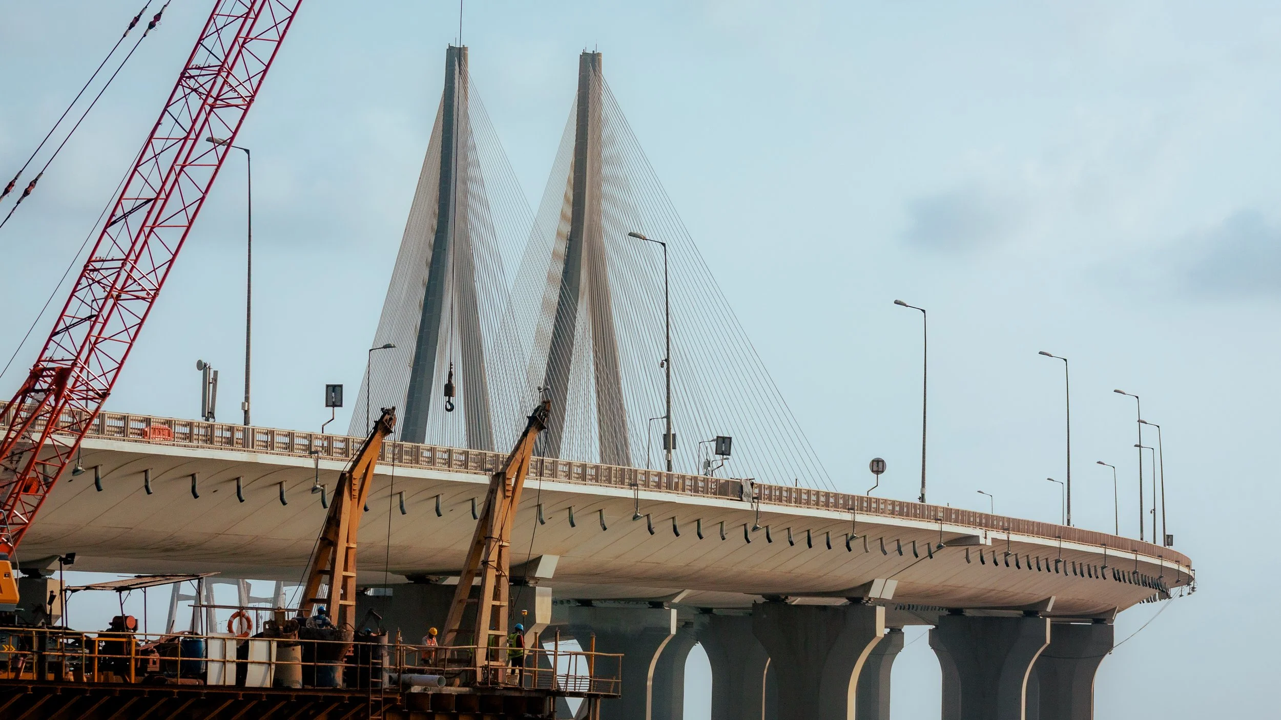 Construction workers on a partially completed cable-stayed bridge with a large red crane nearby, and the bridge's towers and cables visible against a cloudy sky.