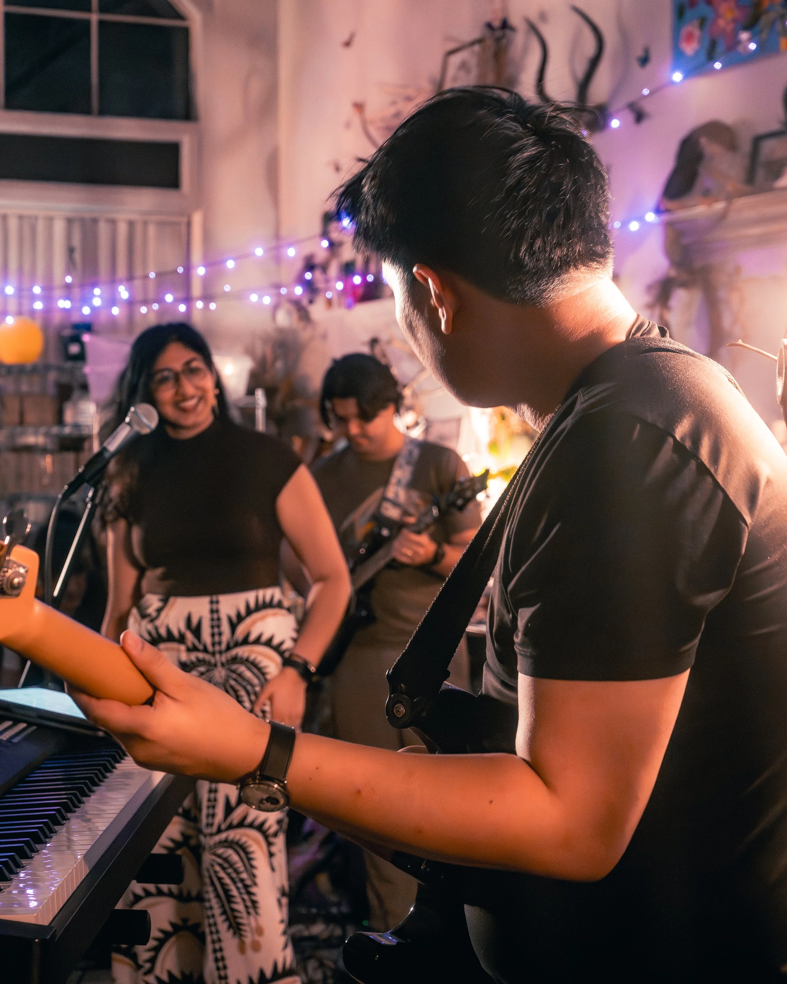 A man playing the guitar and singing at a small gathering with two women, one smiling by a microphone and another playing bass guitar, in a cozy, decorated room with purple string lights.