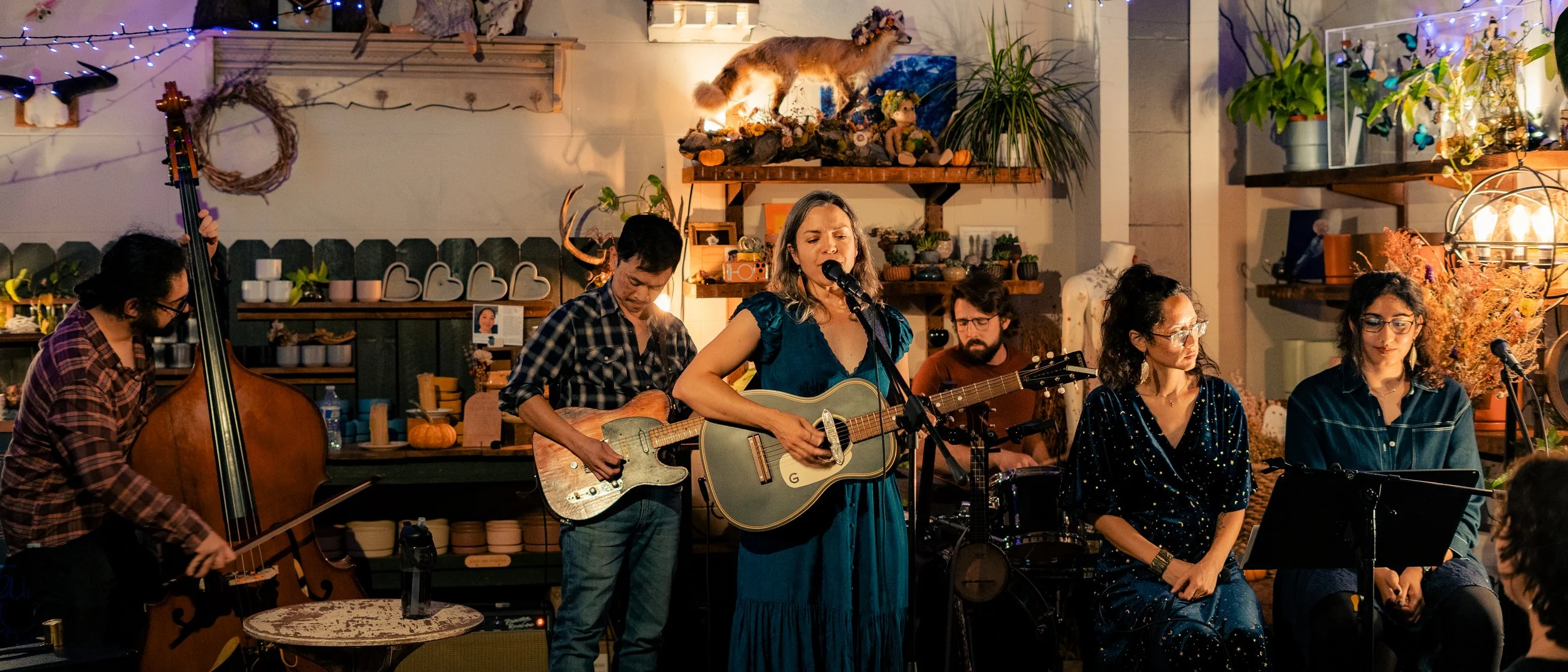 A band performing live music indoors with string lights and decorated plants, with a woman singing and playing guitar at the center.