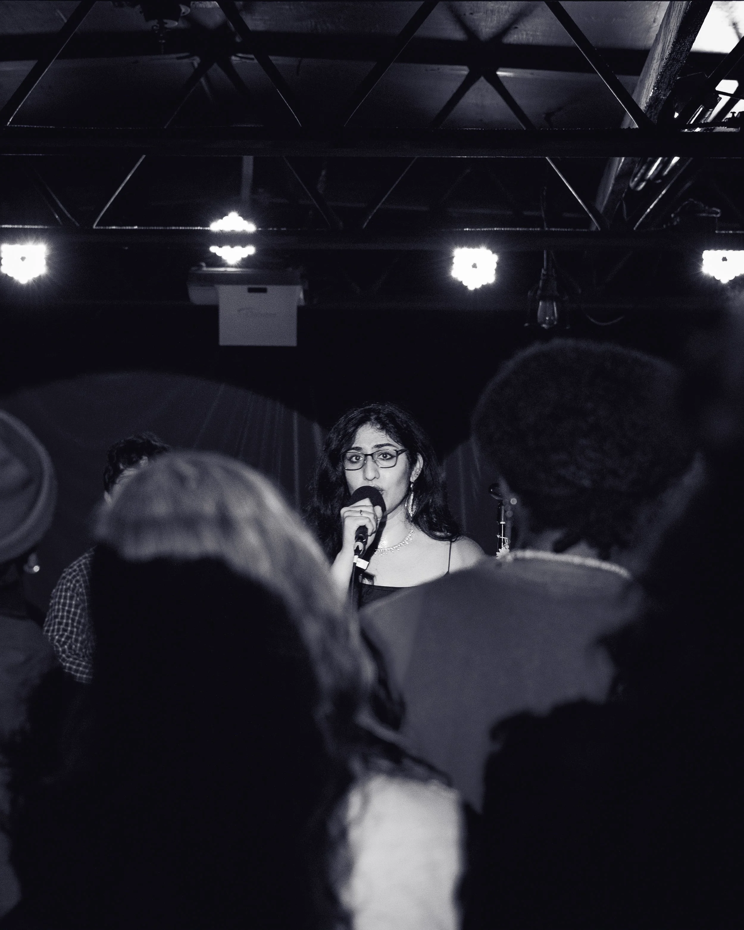 A woman with glasses and long dark hair speaking into a microphone on a stage while an audience listens.
