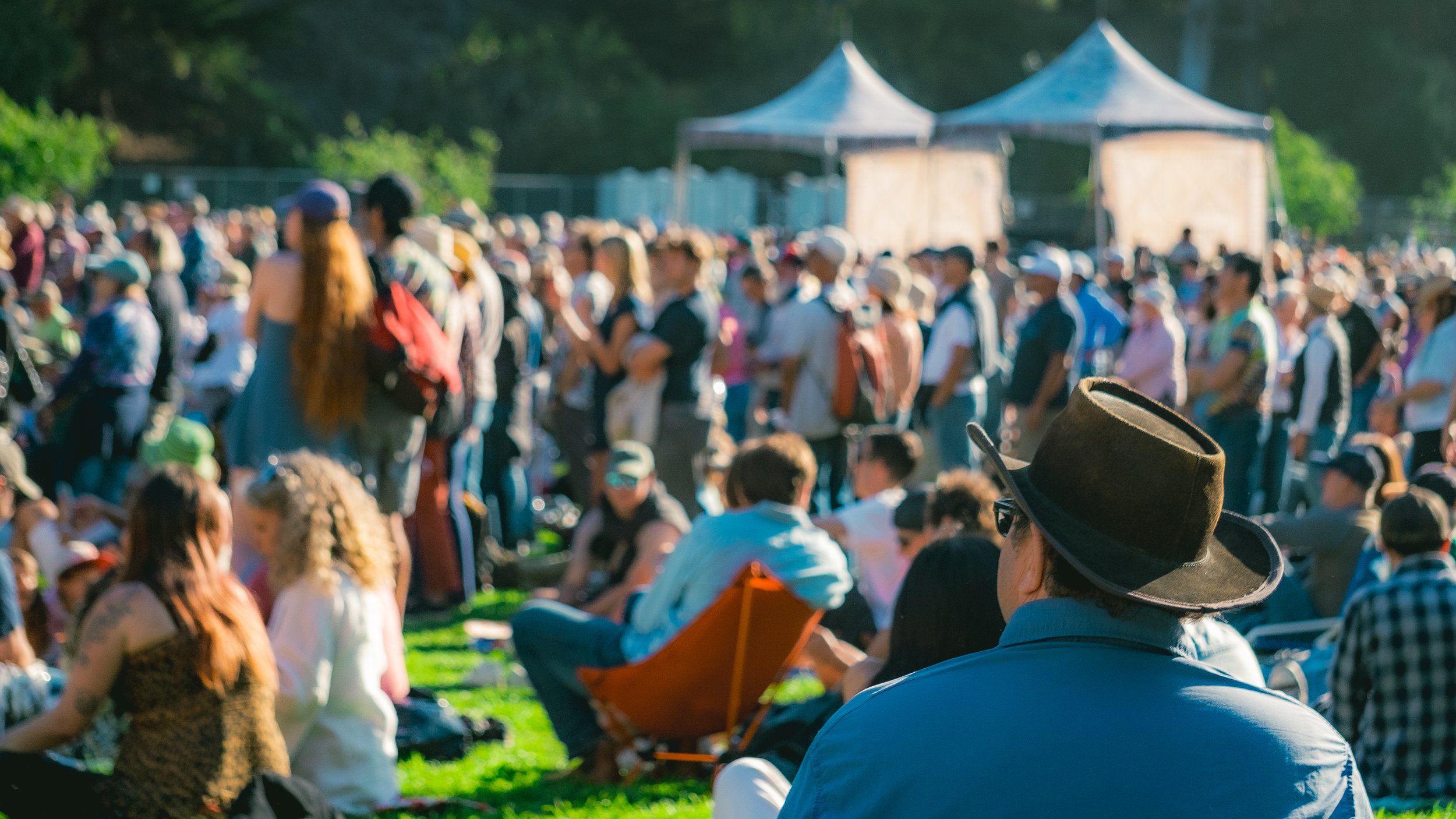 A large outdoor gathering with many people, some sitting on the grass and others standing, under a sunny sky with tents in the background.