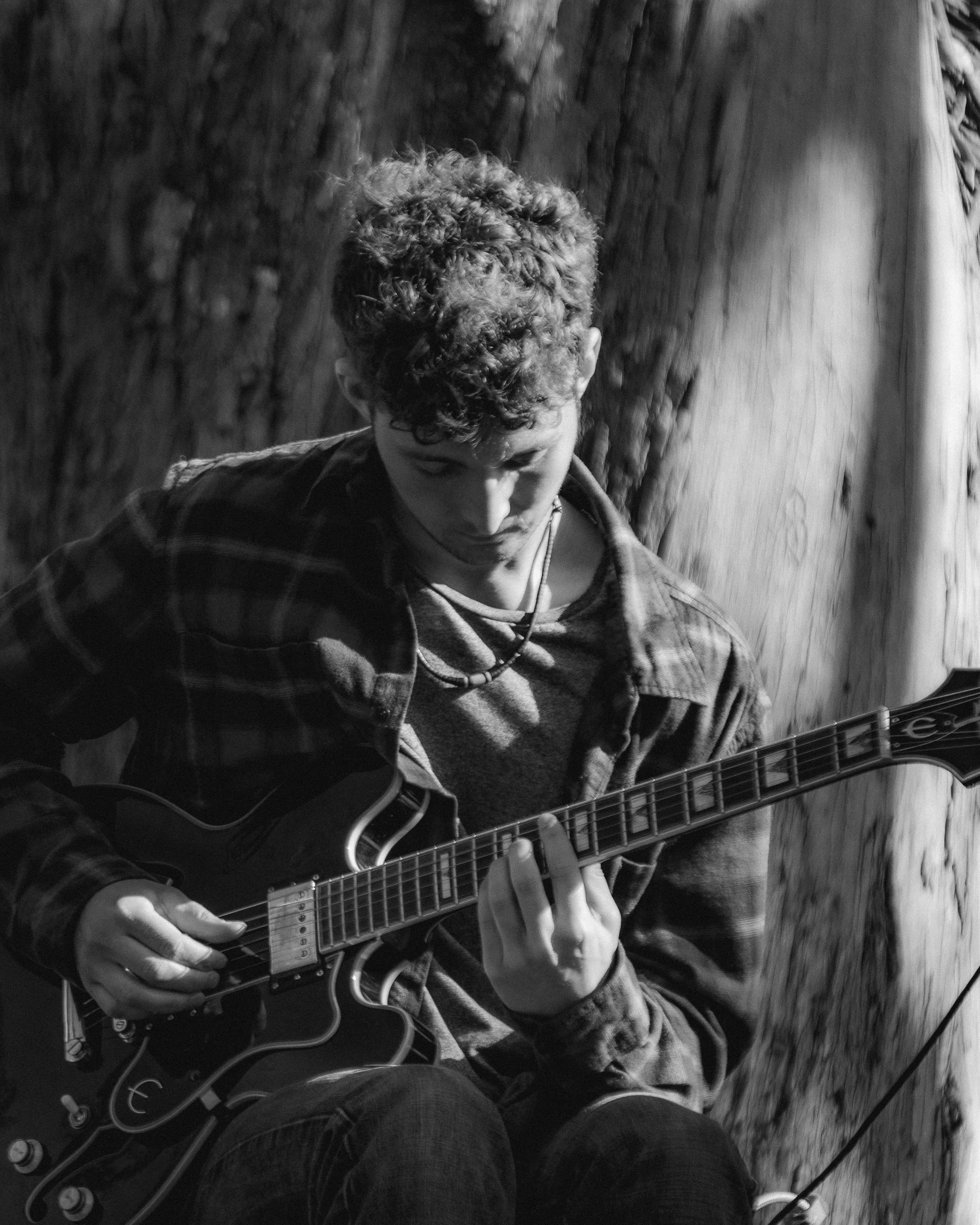 Young man with curly hair playing an electric guitar, sitting against a tree, in black and white.