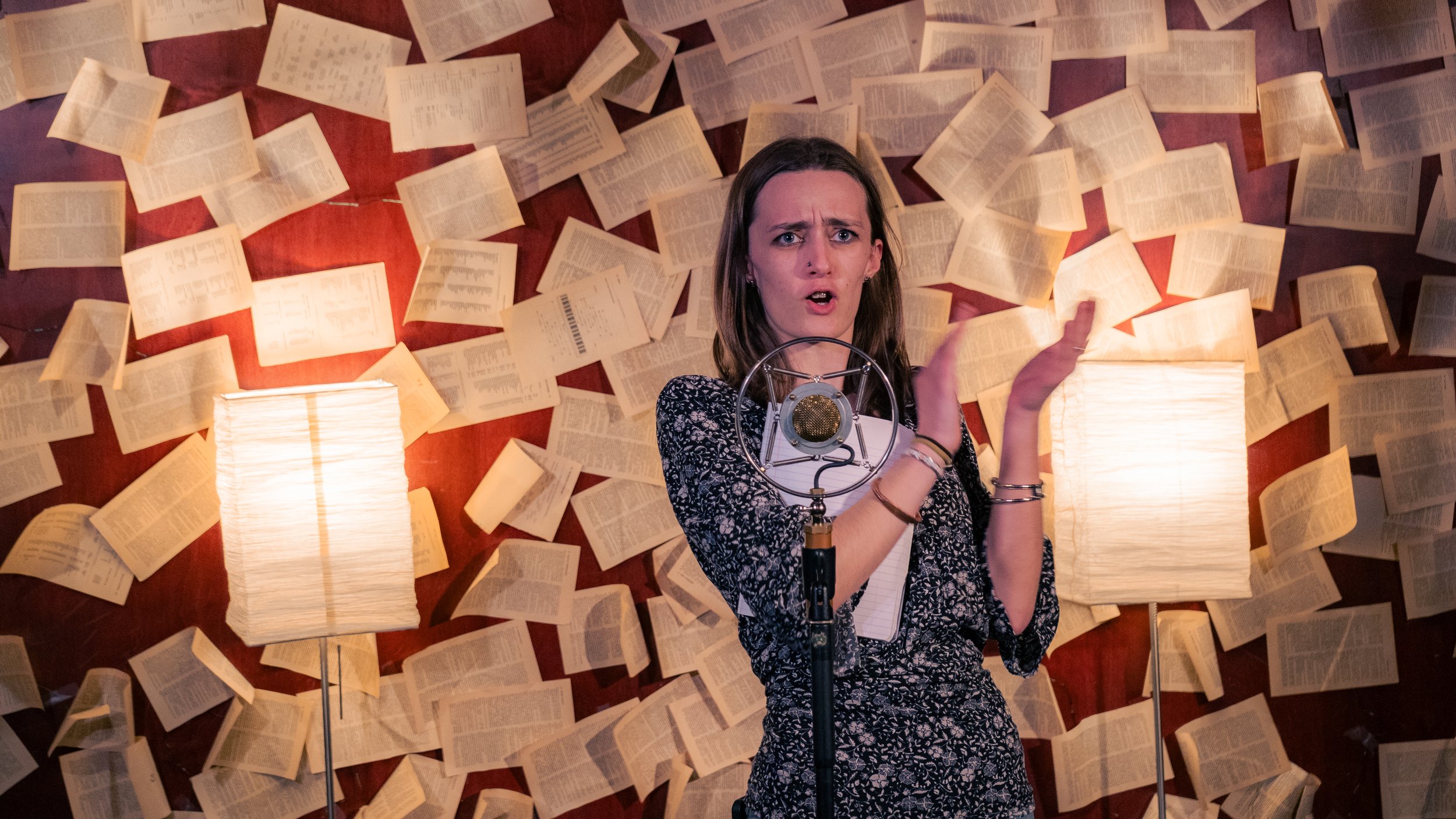 A woman with shoulder-length brown hair standing in front of a microphone, with open books scattered and hung on the red wall behind her, and two lamps on either side illuminating the scene.