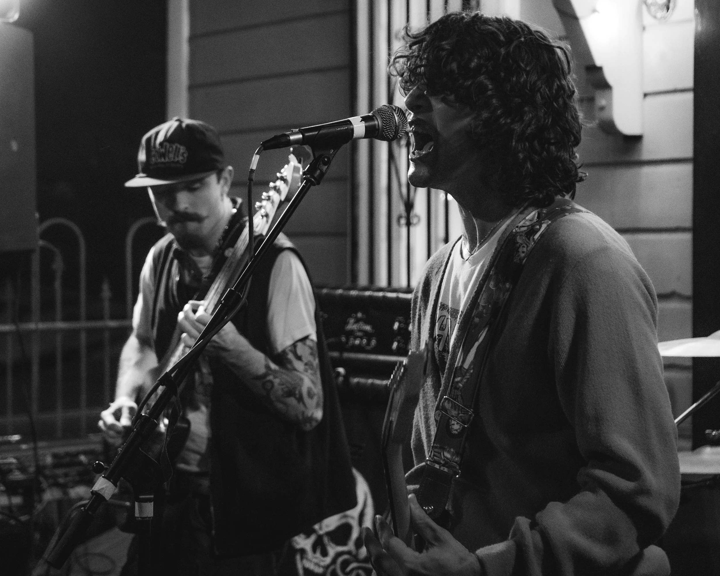 Two musicians performing indoors. The one on the right is singing into a microphone, playing an electric guitar, wearing a hoodie. The other on the left is playing an electric guitar, wearing a baseball cap and tattoo sleeves. The scene is in black a