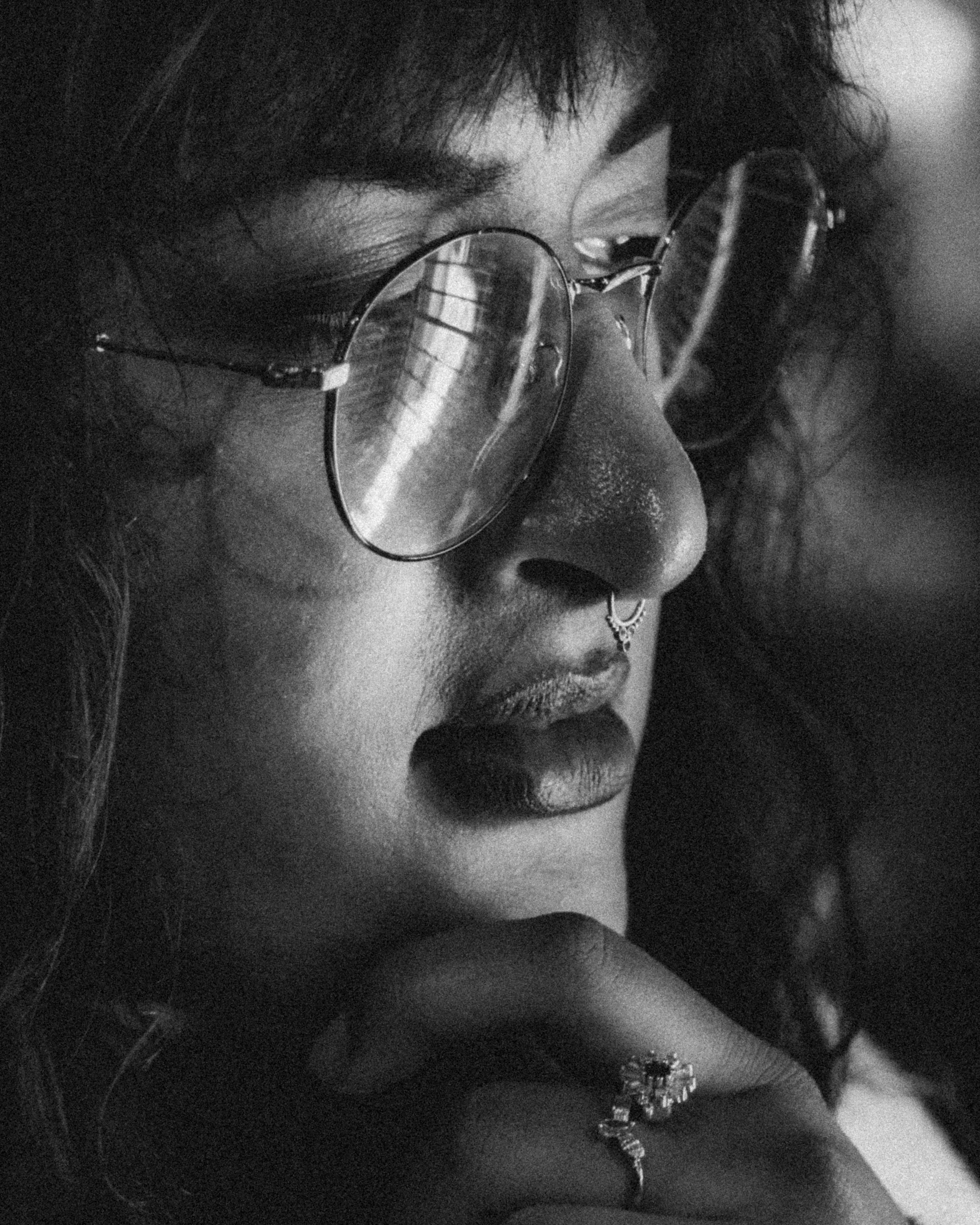 A close-up black and white photo of a woman wearing round glasses with reflections, a septum piercing, lipstick, and jewelry. She is resting her face on her hand.