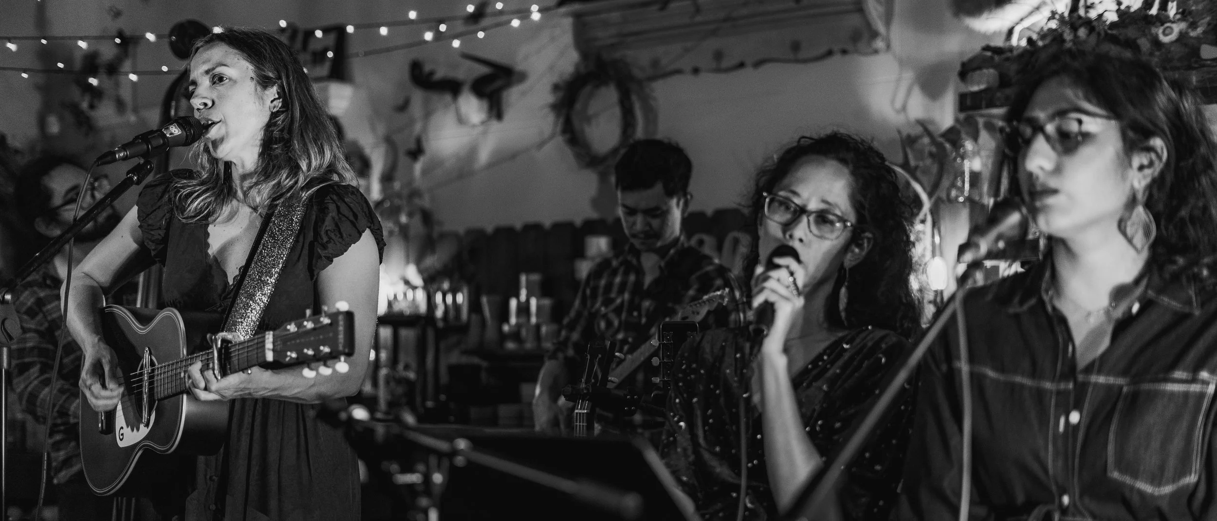 Black and white photo of four women performing music: the woman on the left is singing and playing guitar, the second is singing into a microphone, and the third is also singing into a microphone. The background shows a cozy indoor setting with strin