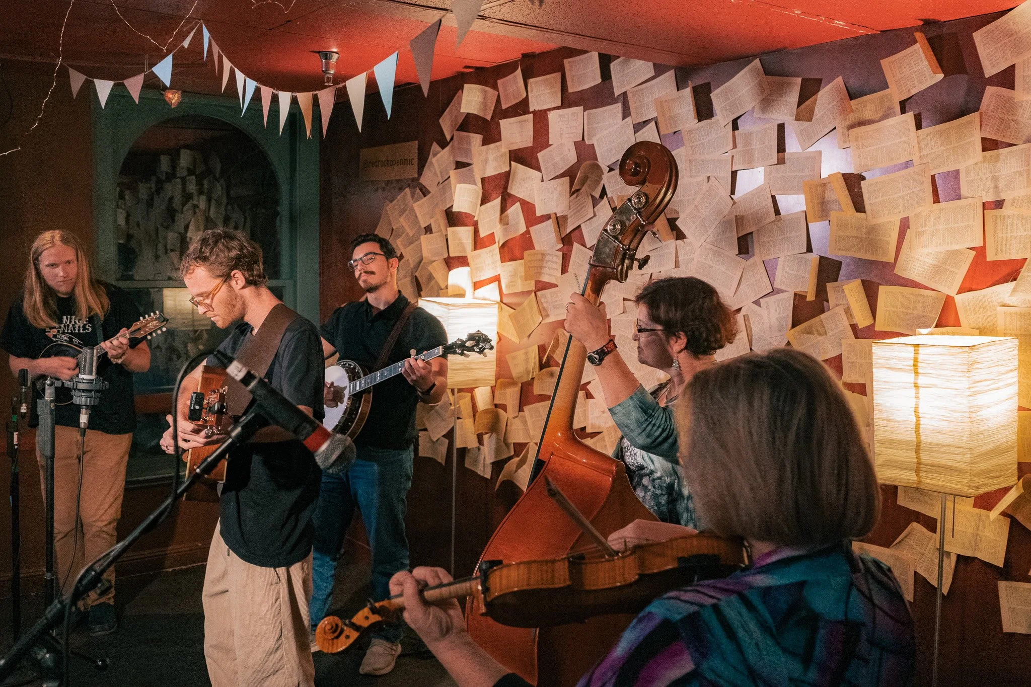 A group of musicians performing in a cozy, decorated room with paper pages on the wall and soft lighting.