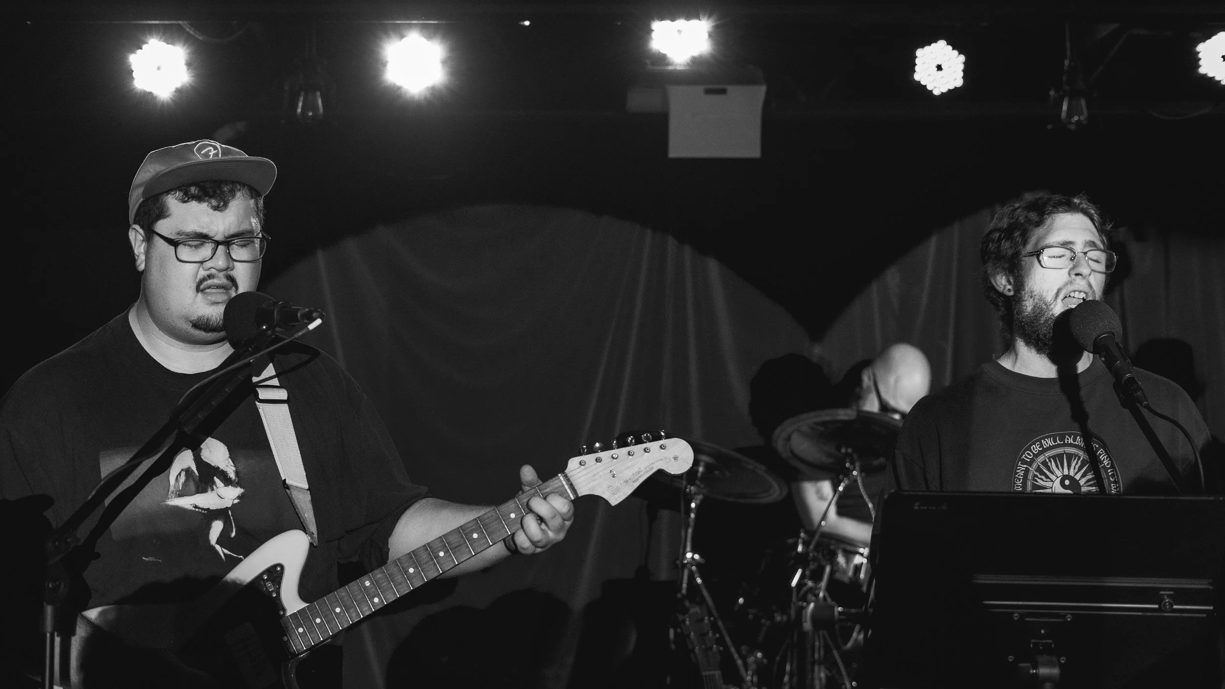 Two men performing music on stage with microphones, one playing an electric guitar and the other singing, with a drum set in the background, in a dimly lit setting.