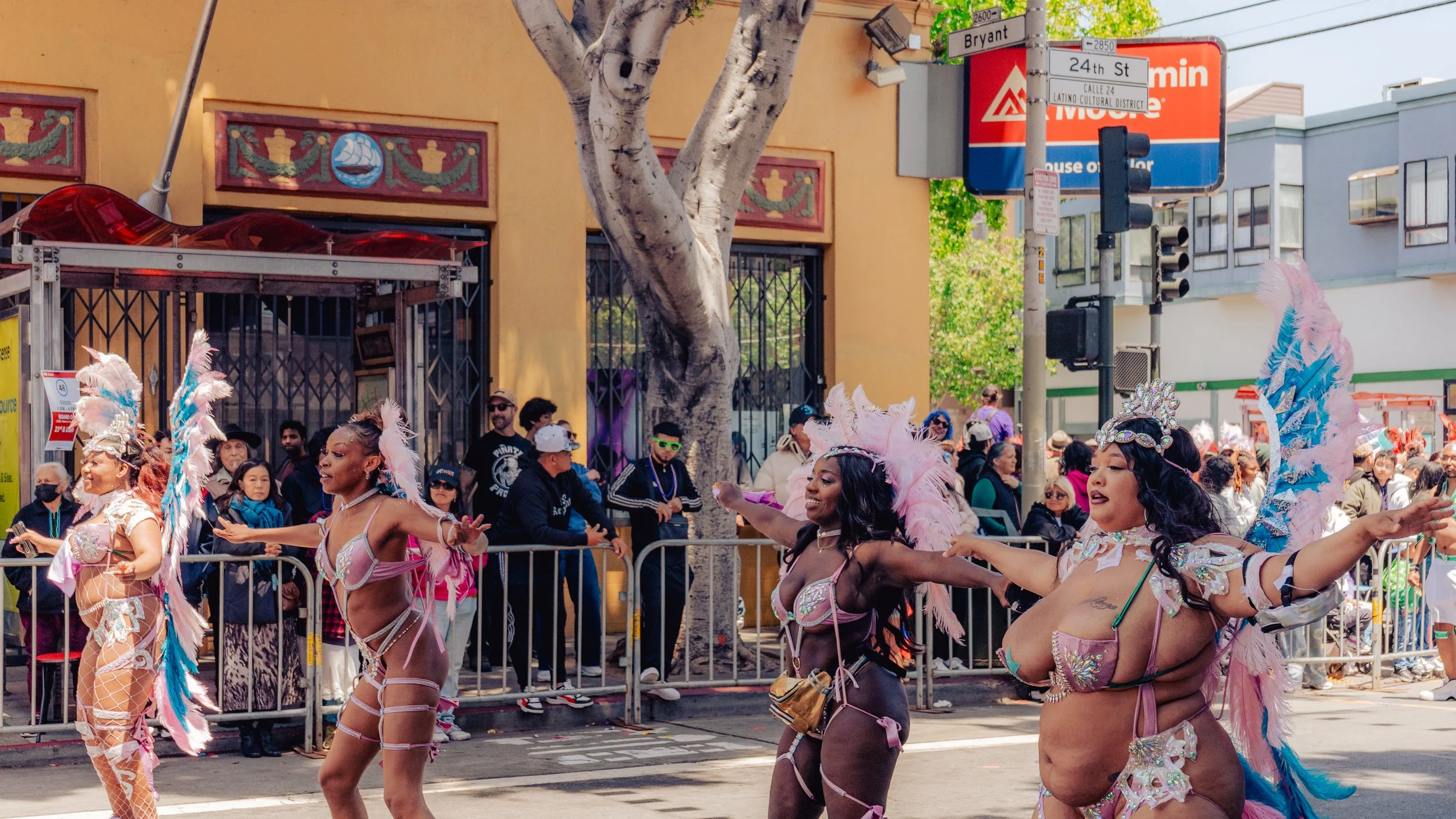 Four women in colorful costumes with feathers and decorative accessories dancing in a parade on a city street, with spectators watching behind them.