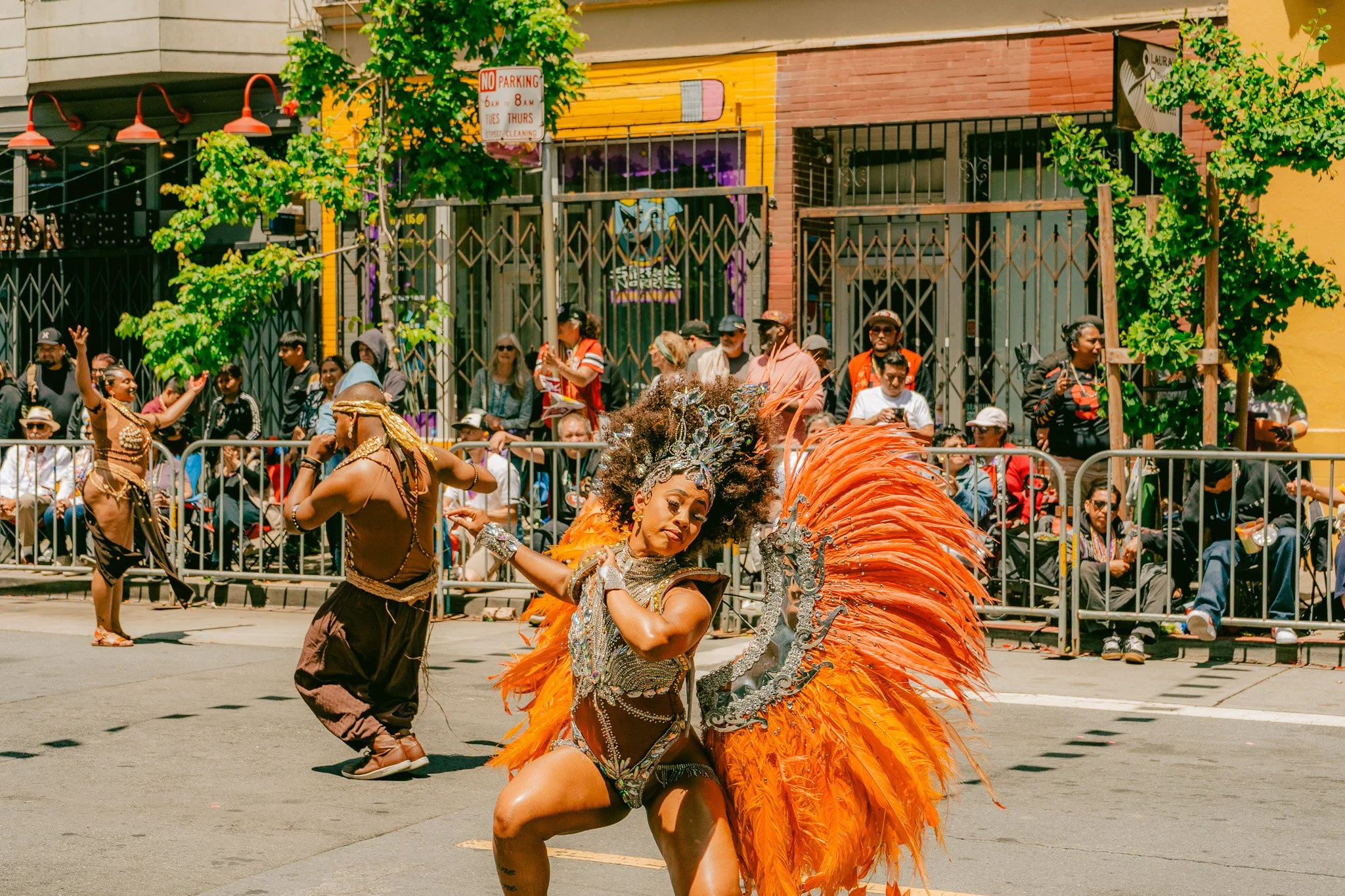 A woman in a colorful carnival costume with large orange feathers dancing in the street during a parade, with spectators watching behind a metal barricade.
