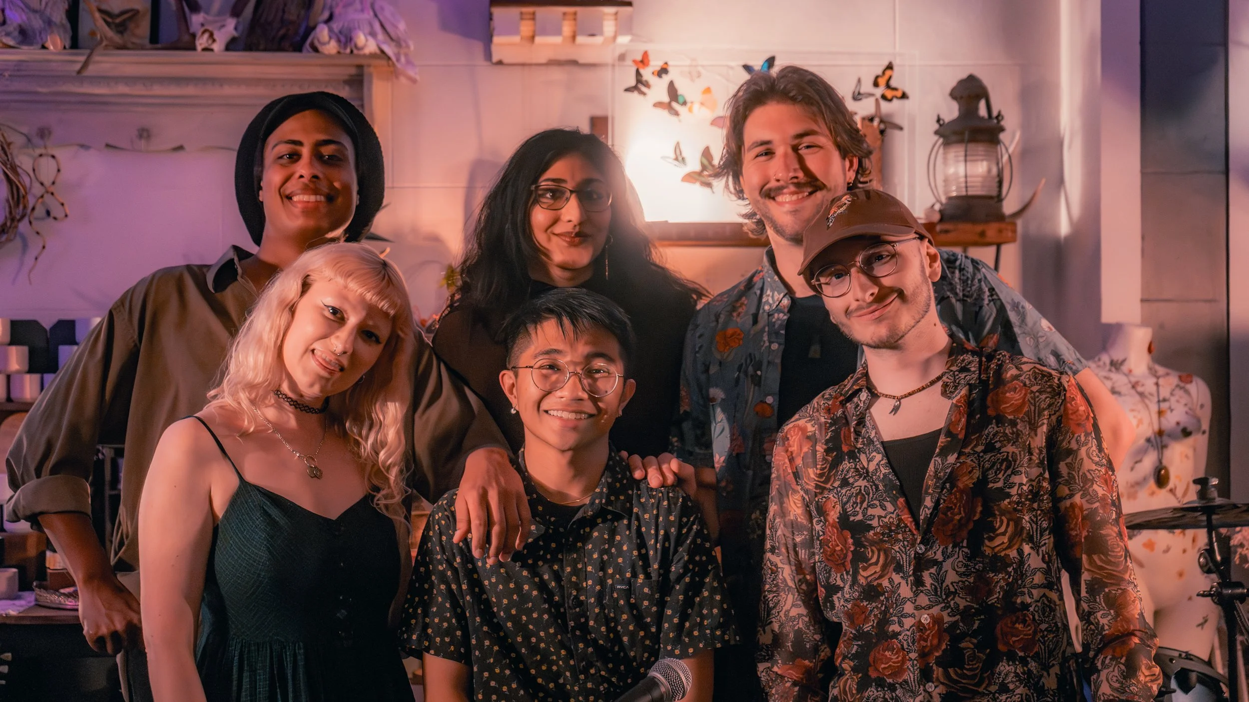 Group of seven diverse young adults posing together in a room decorated with butterfly artwork and warm lighting, smiling at the camera.