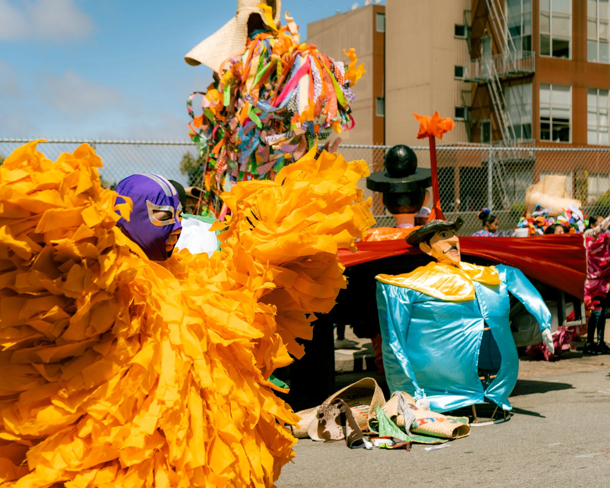 Colorful parade costumes and masks on display with buildings and a chain-link fence in the background.