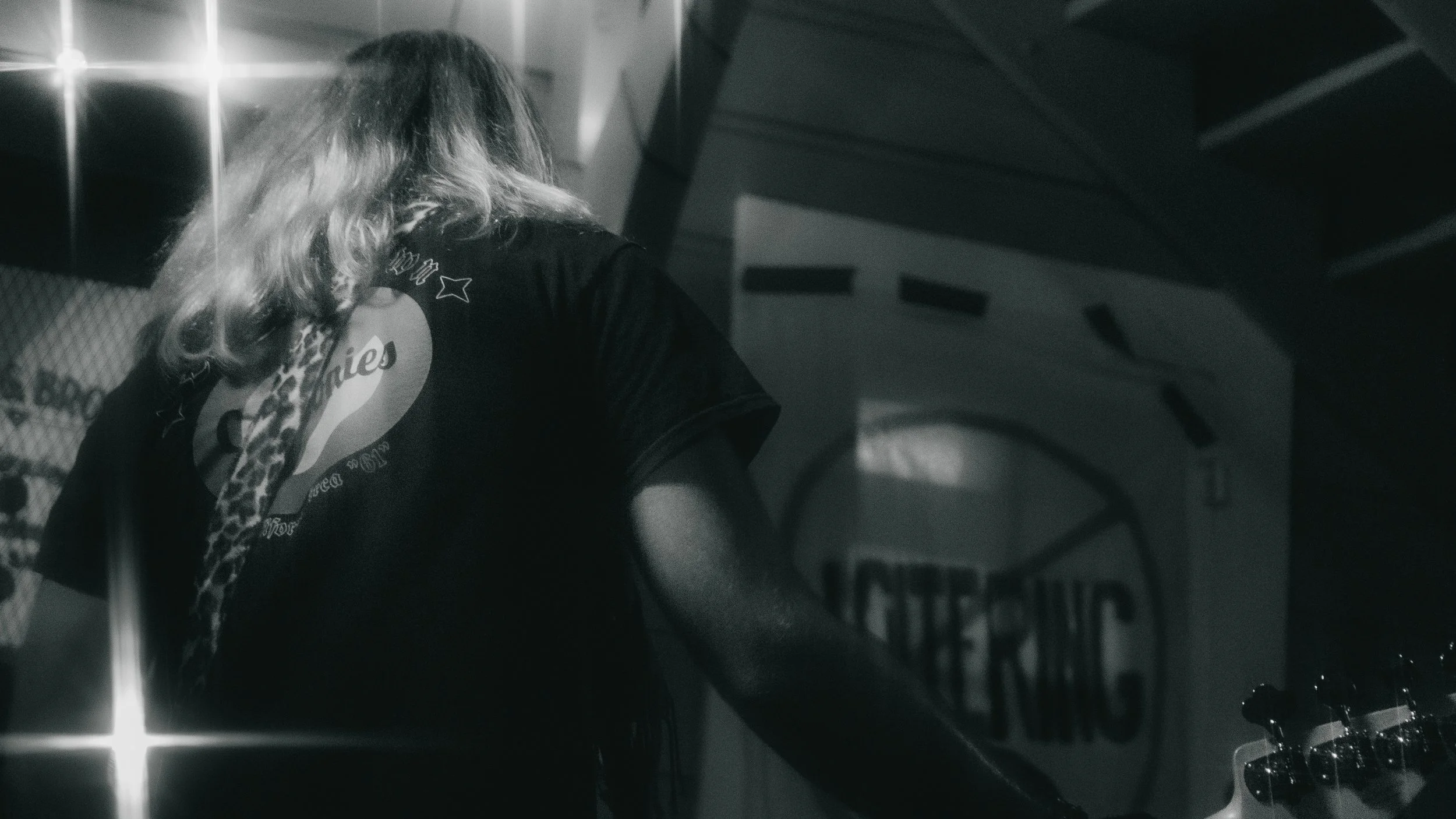 A person with long, wavy hair wearing a black T-shirt, seen from behind, in what appears to be a music or event venue, with a large sign in the background that reads 'INTERNS' and some equipment visible to the right.