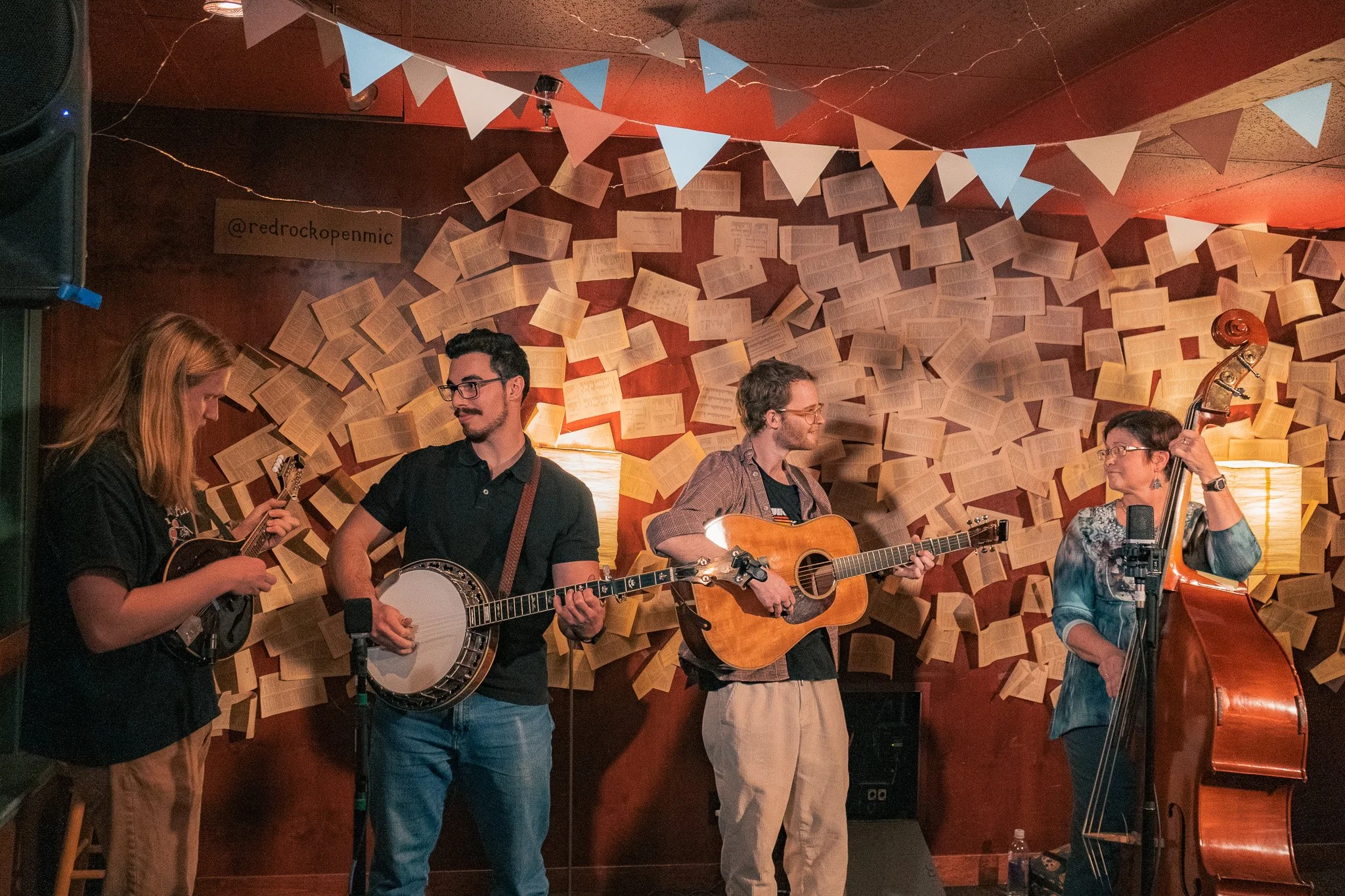 Four musicians playing instruments on stage with a background of floating book pages and triangular banners.