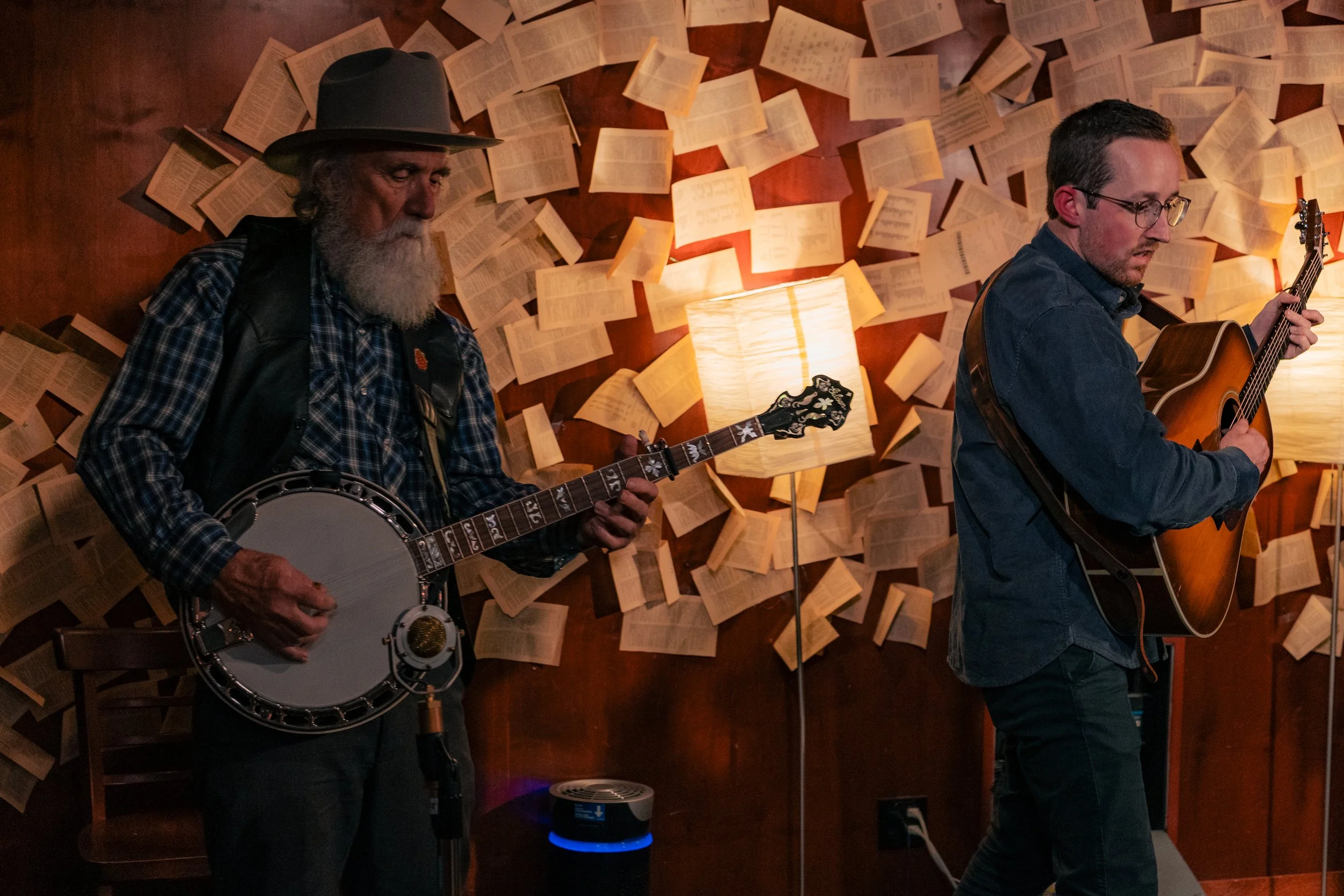Two men playing guitars and a banjo in a room with a wall covered in open books and a lamp.