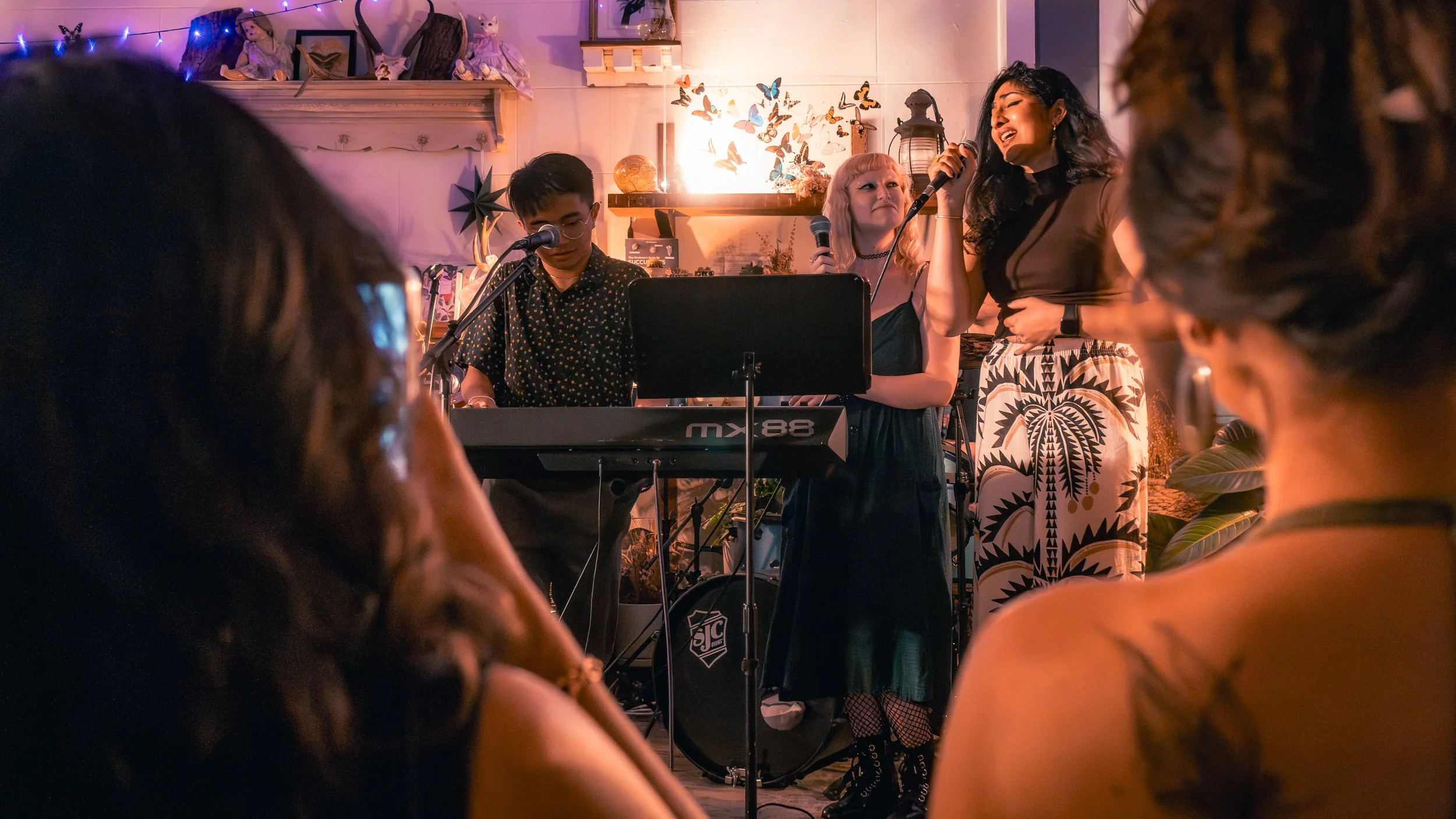 A musical performance with three singers and a keyboard player, set in a cozy, decorated indoor space with members of an audience watching.
