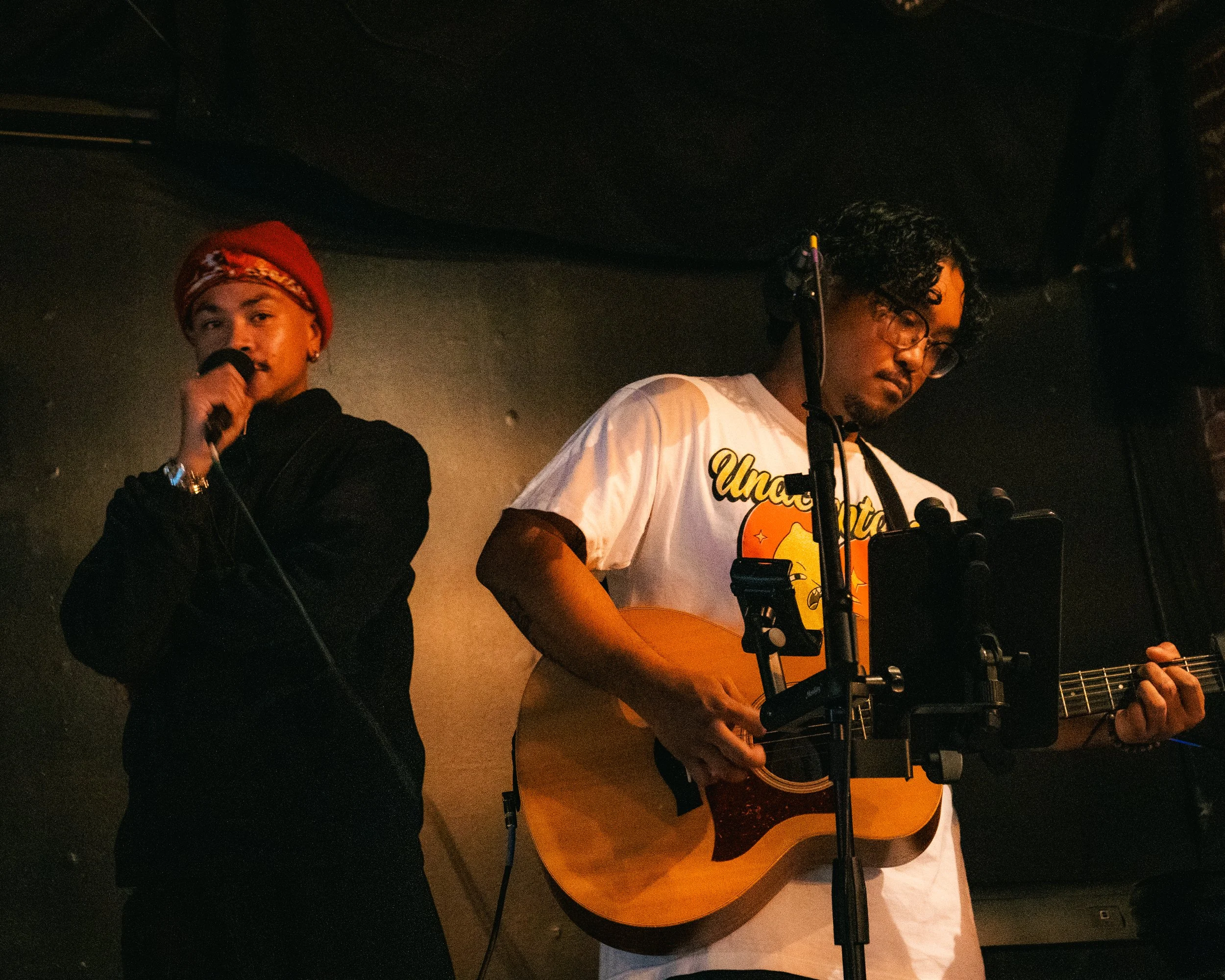 Two musicians perform on stage, one singing into a microphone and wearing a red beanie, and the other playing an acoustic guitar and wearing glasses and a white T-shirt.