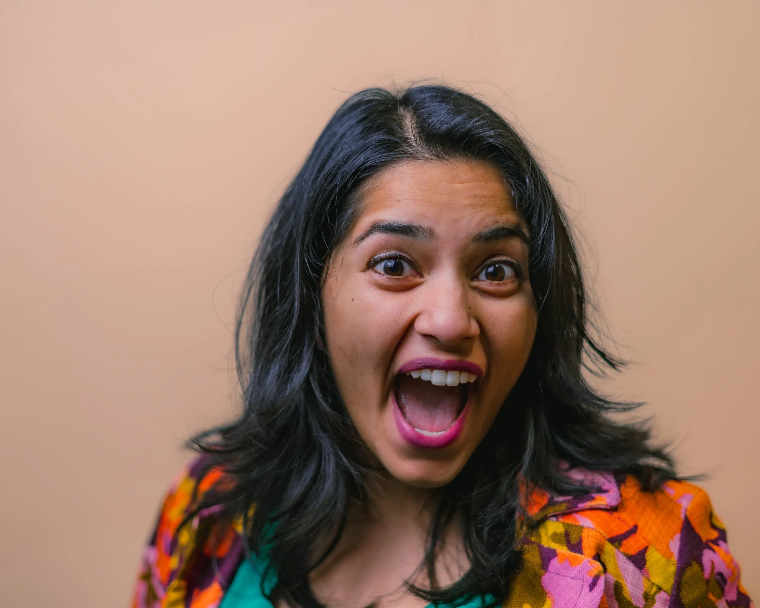 A woman with dark hair and an excited expression, mouth open wide, wearing a colorful patterned jacket over a green top.