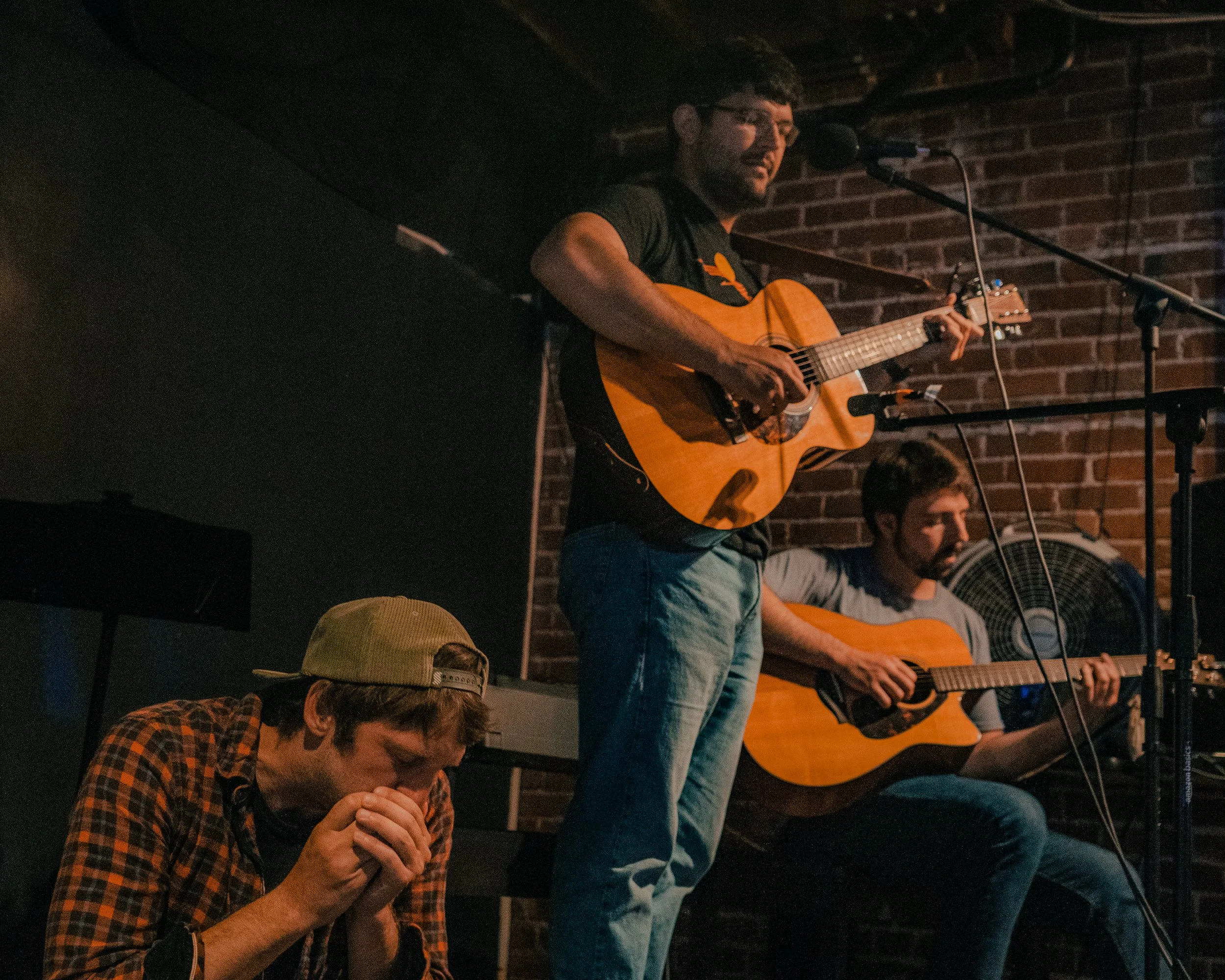 A group of four people, with three playing guitars and one kneeling with eyes closed, performing on a stage with brick wall background in a dimly lit venue.