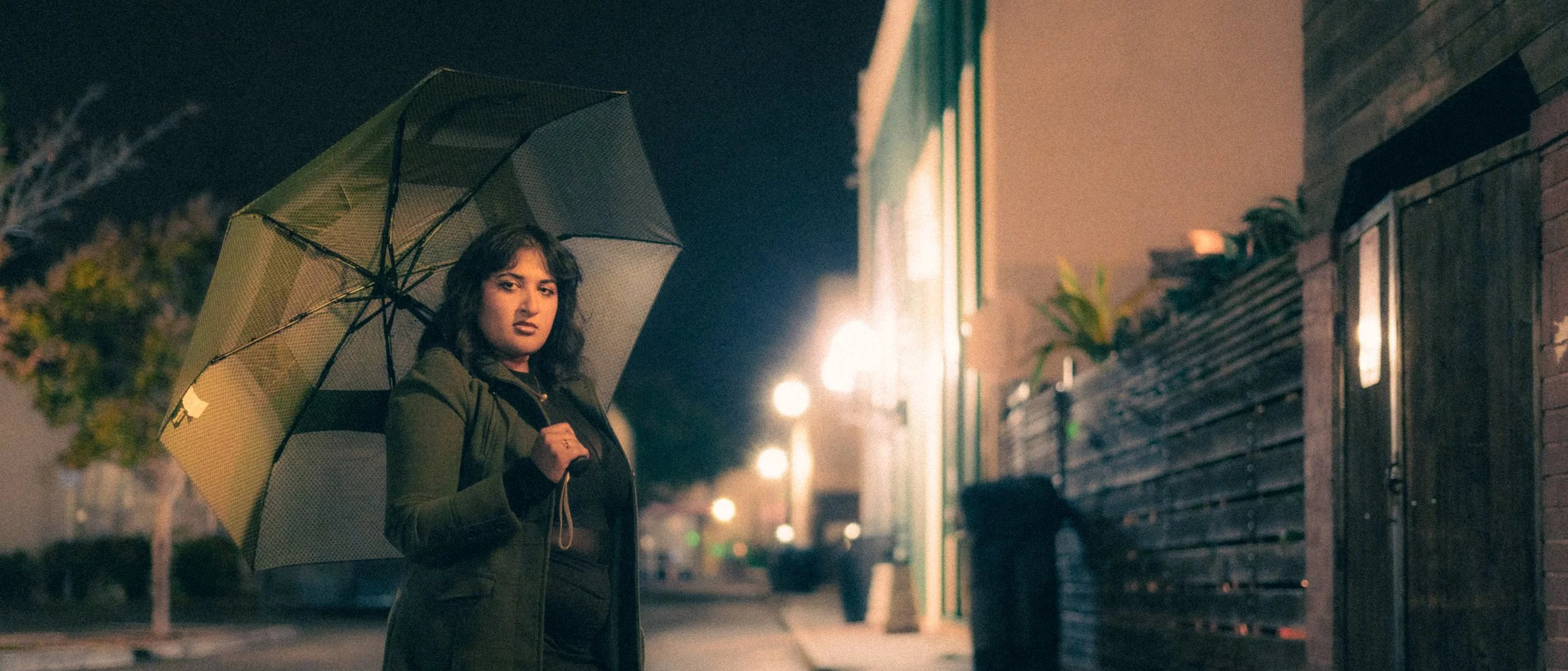 A woman standing on a city street at night, holding an umbrella. The scene is illuminated by streetlights, with a building and trees in the background.
