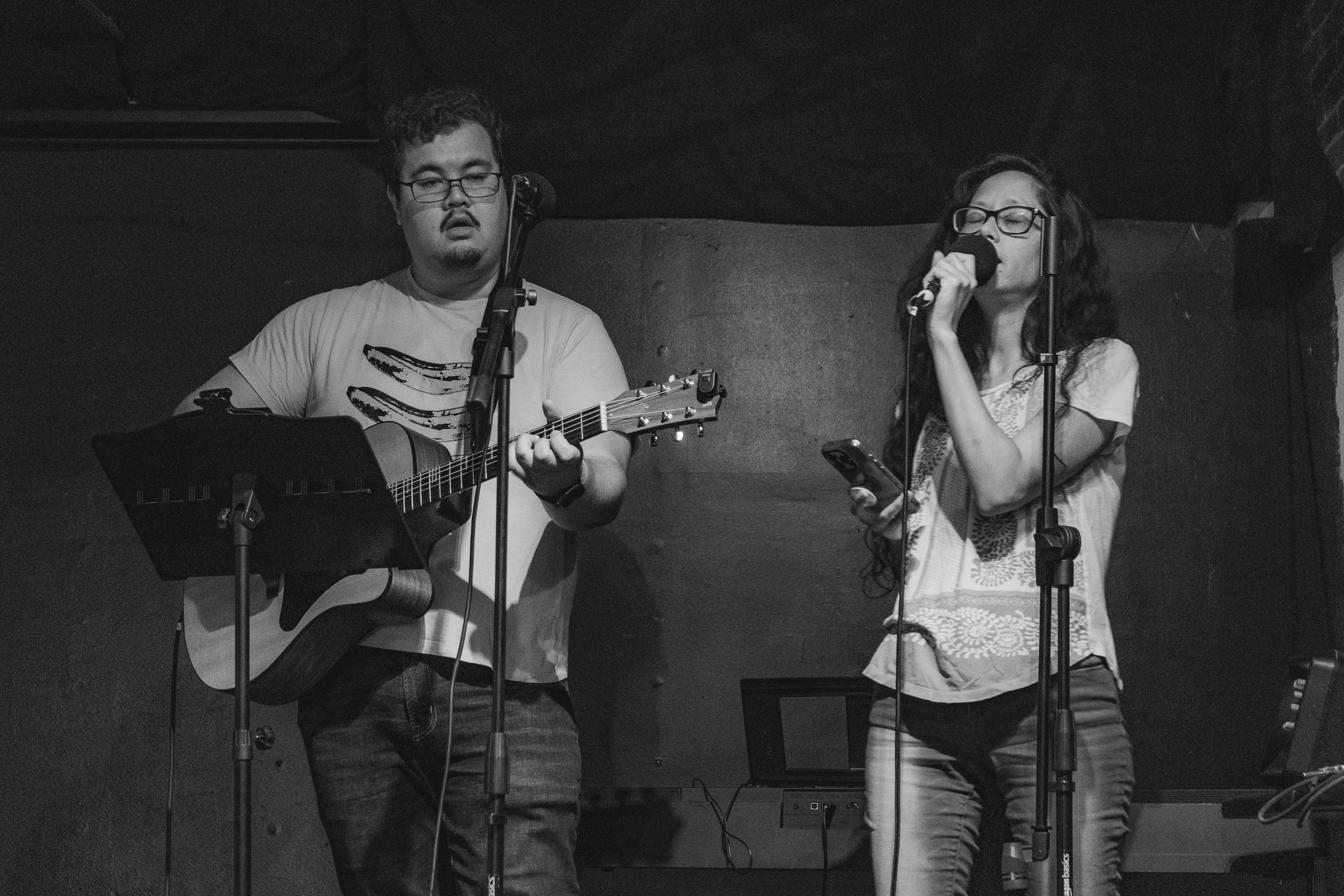 A man playing guitar and a woman singing into a microphone on stage in a dark setting.