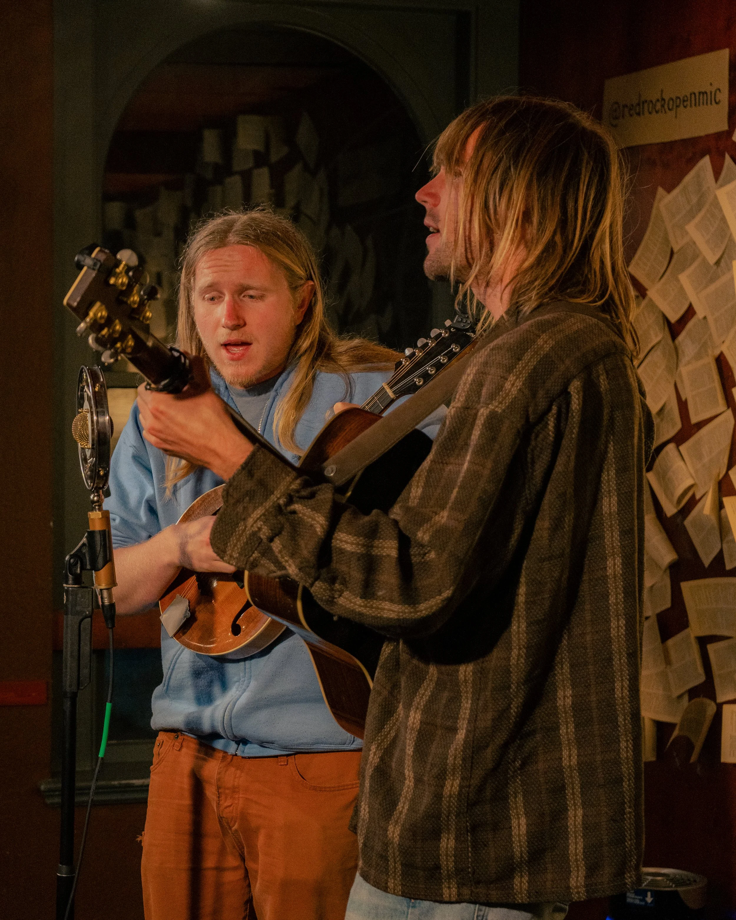 Two men singing and playing guitars in a cozy, dimly lit room, with sheet music or pages pinned on the wall behind them.