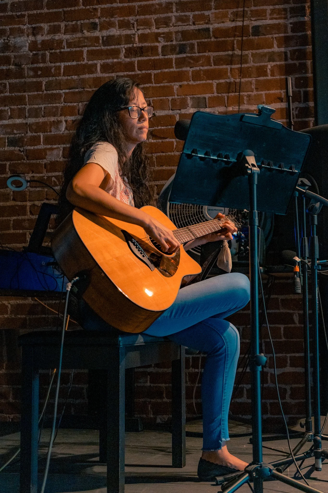 A woman with long dark hair and glasses playing an acoustic guitar on a small stage with a brick wall background.
