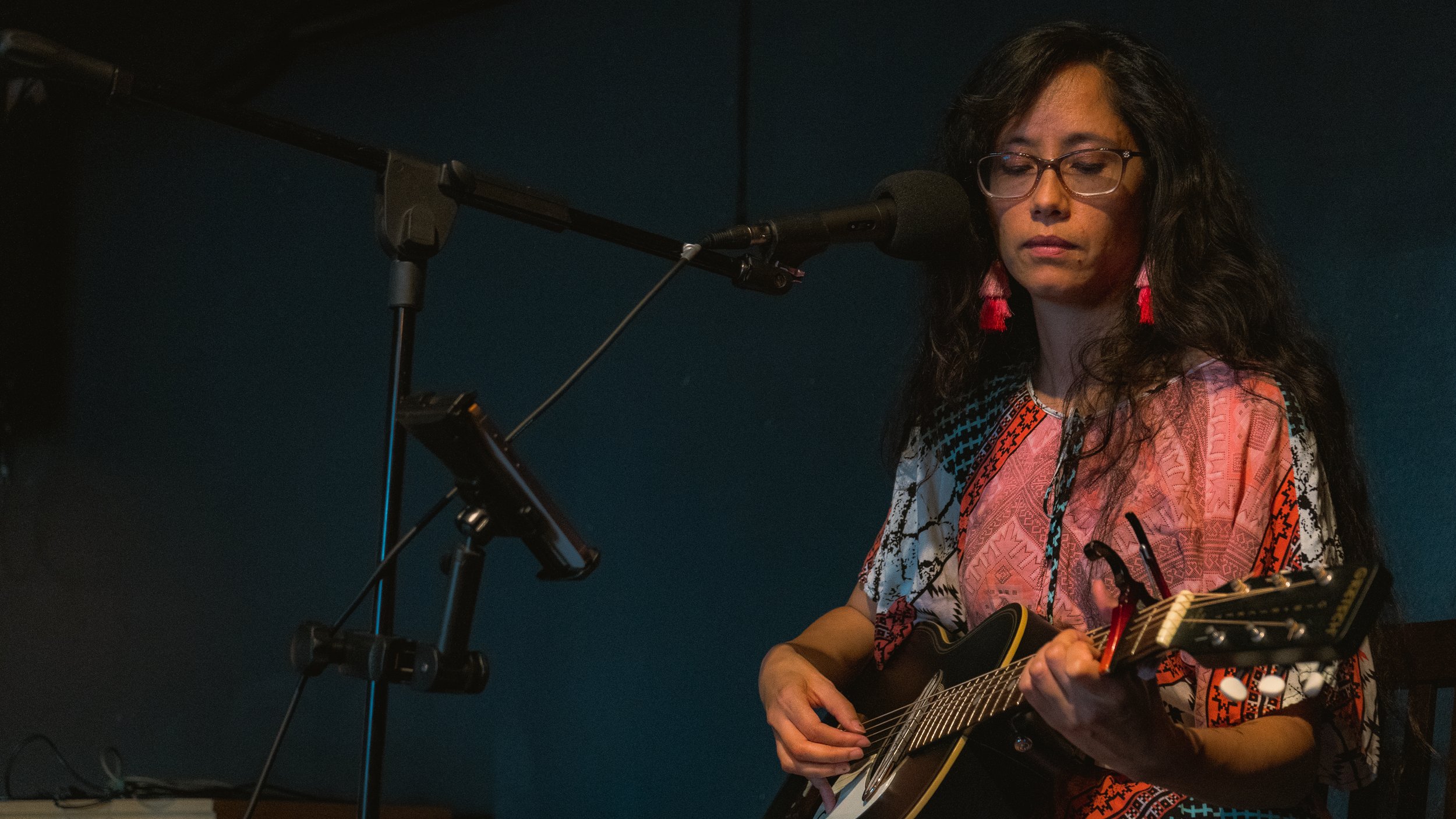A woman with glasses and long dark hair playing an acoustic guitar in front of a microphone on a stand indoors.