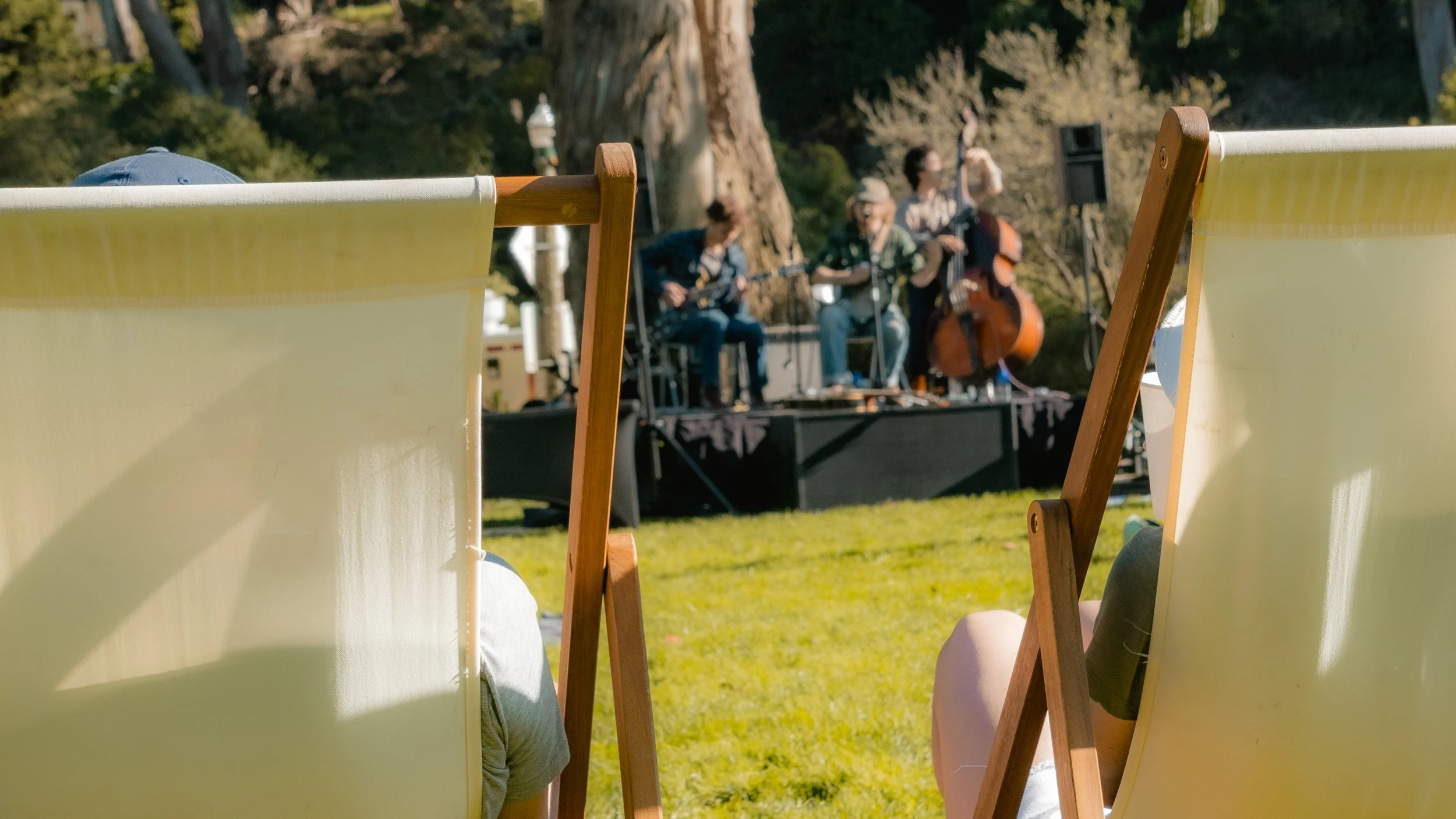 An outdoor scene with a small stage where a band of three musicians is playing. The band includes a guitarist, a bassist, and a drummer. The audience is sitting on lawn chairs, with two yellow chairs visible in the foreground, facing the stage. The s