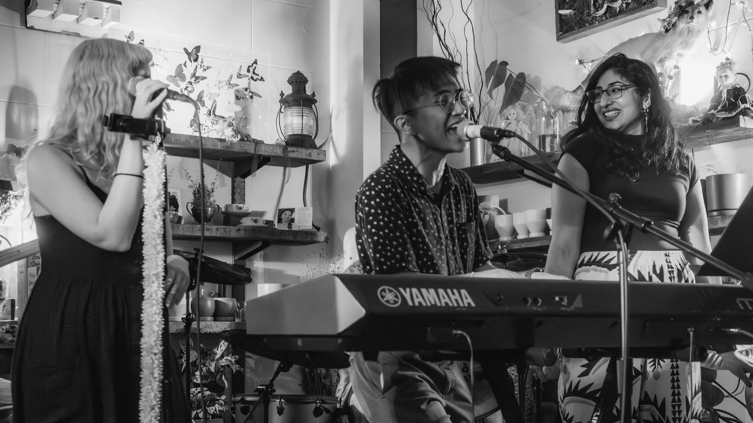 Three women singing and playing keyboard at an indoor gathering with decorative items and fairy lights in the background.