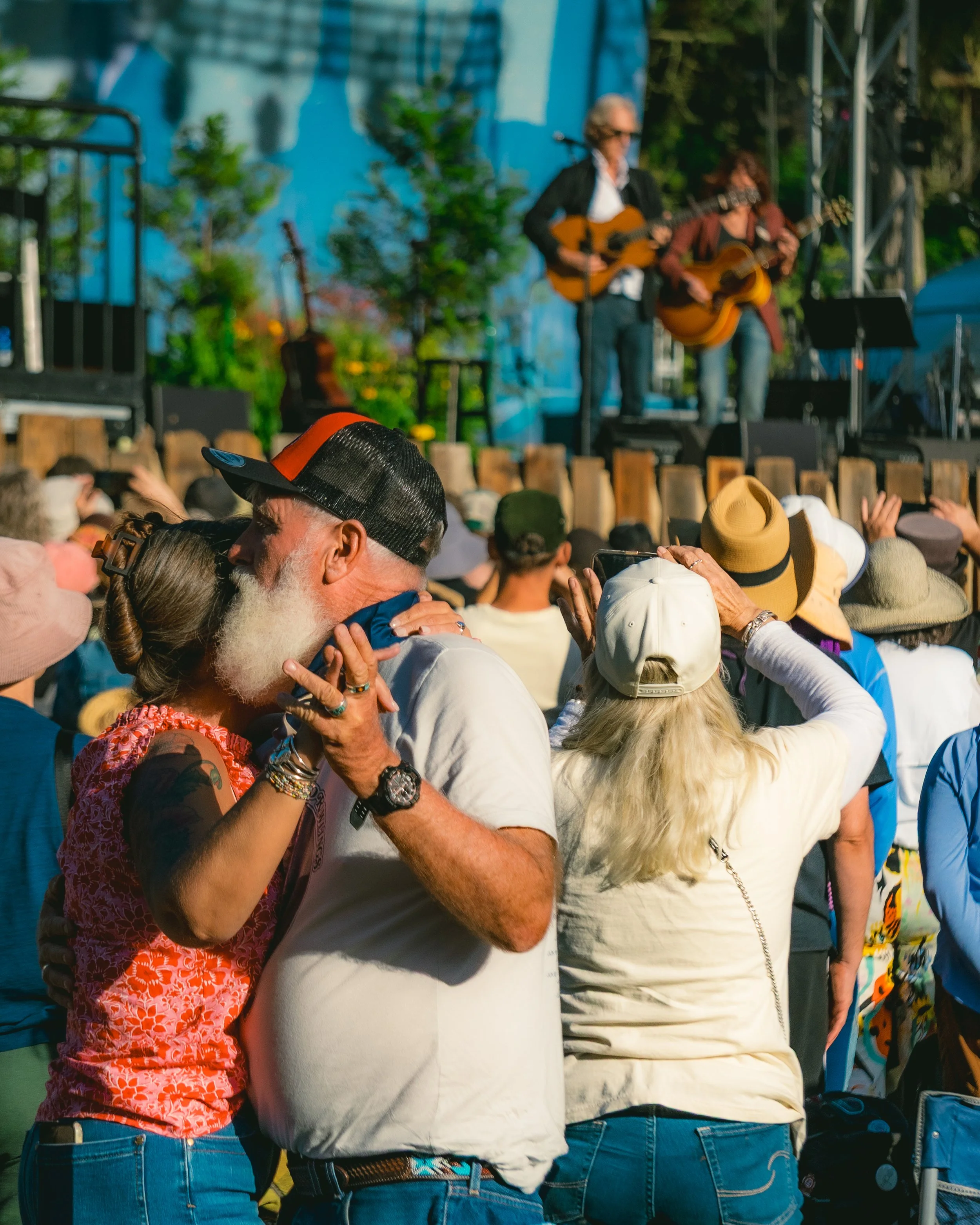 People at an outdoor concert, a man and woman hugging while dancing, with a band playing on stage in the background.