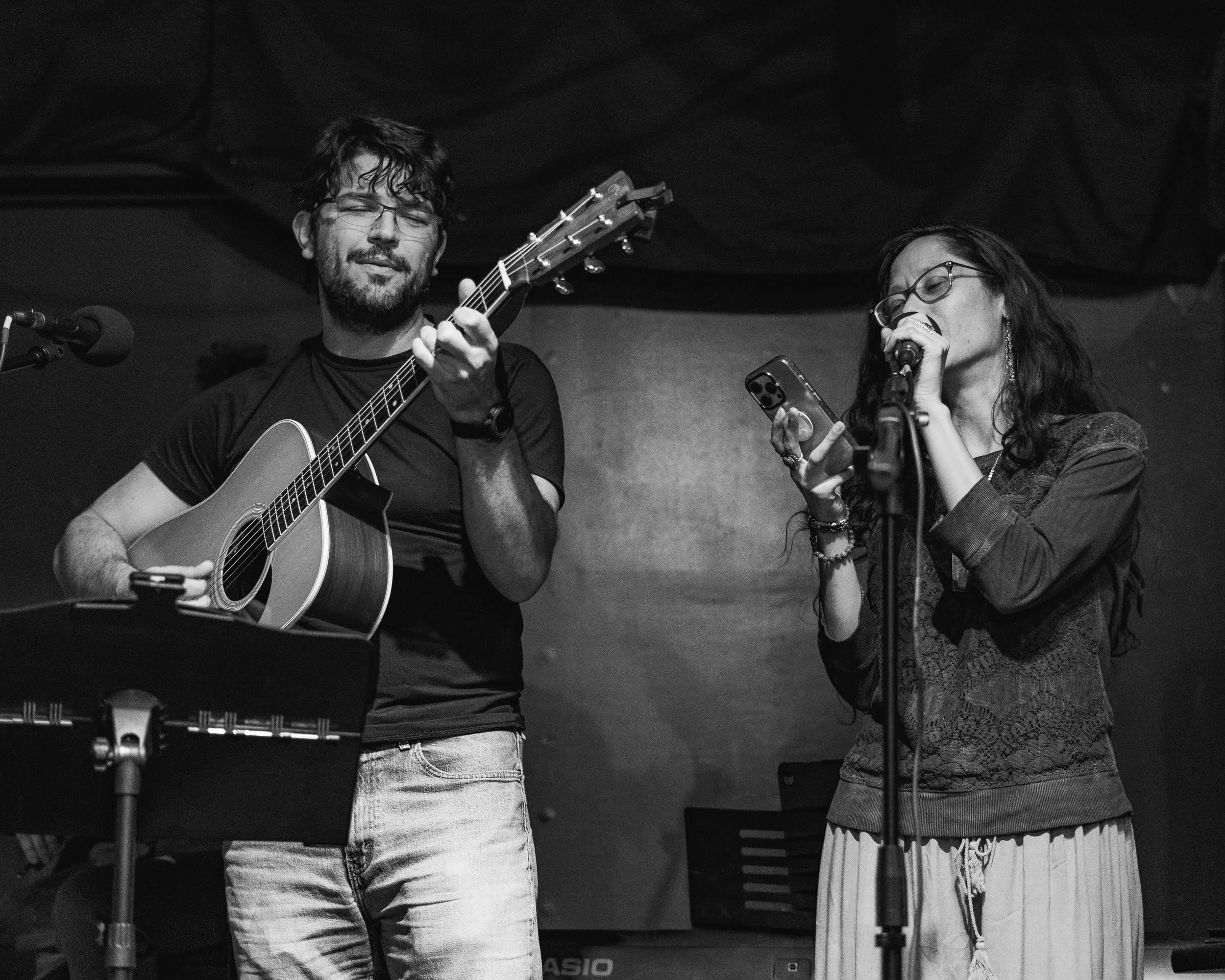 A man with glasses and a beard playing an acoustic guitar and a woman with glasses singing into a microphone; both are on stage with musical equipment in the background.