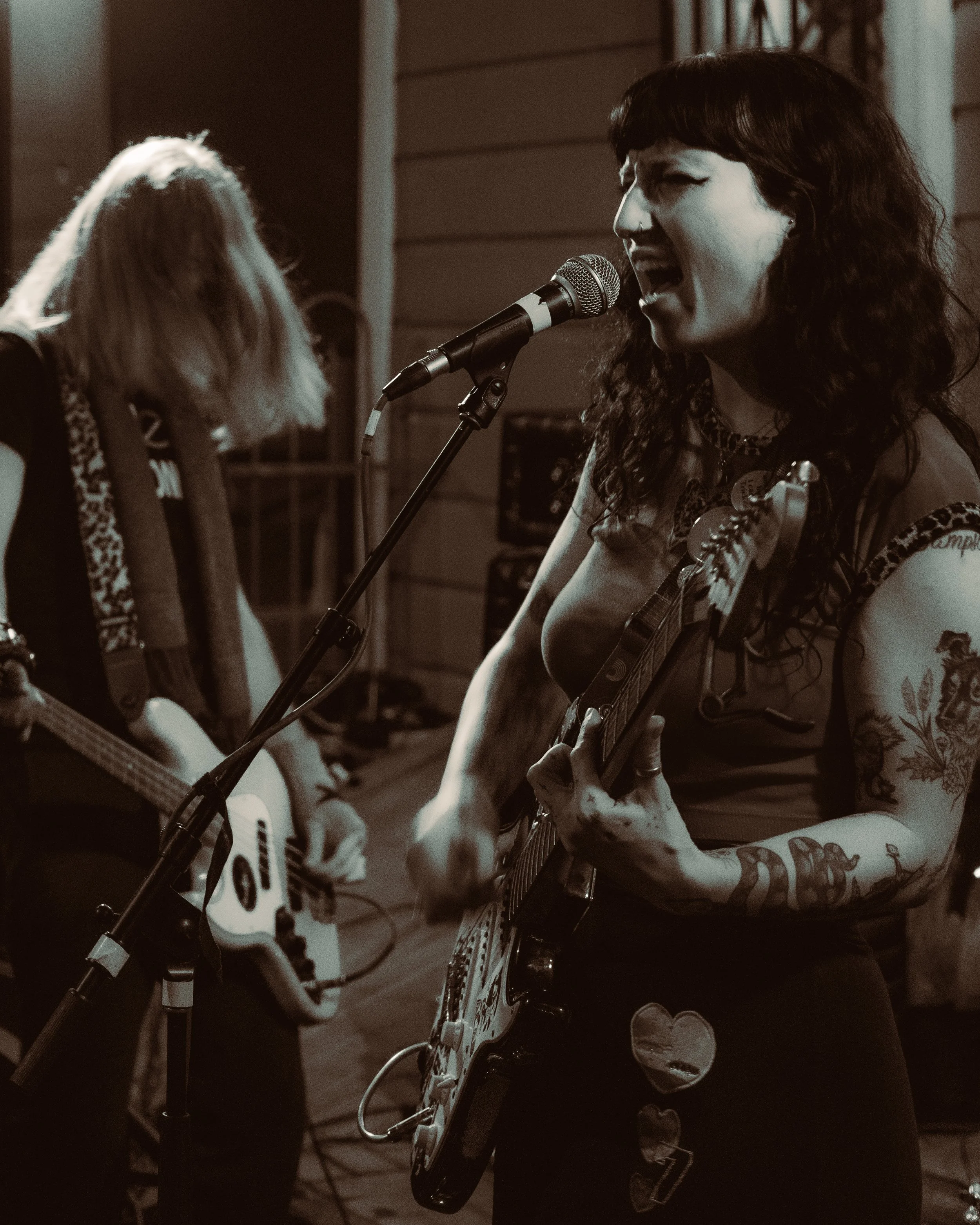 A woman with dark, curly hair singing into a microphone and playing an electric guitar, with another woman in the background playing an acoustic guitar, performing together.