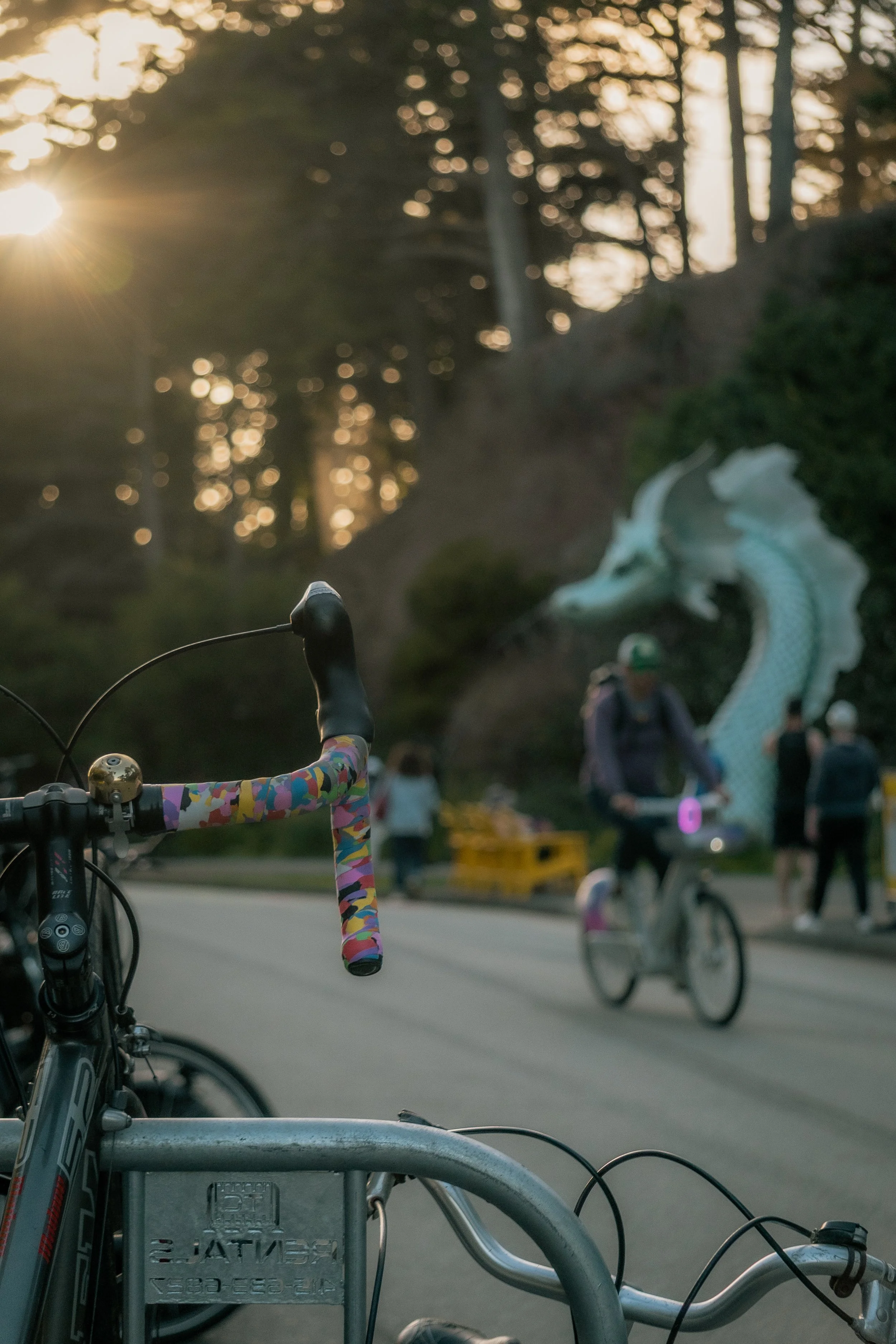 Bicycle handlebar with colorful tape, with a dragon sculpture and people riding bikes in the background during sunset.